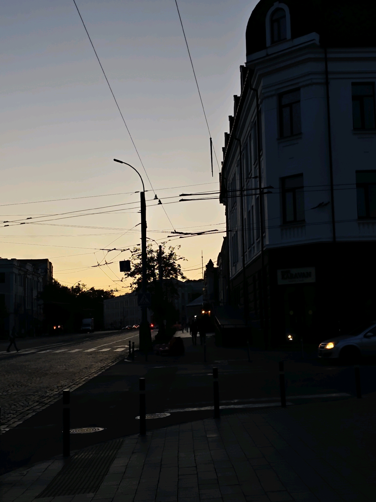 The image depicts a street scene in Vinnytsia, Ukraine, during twilight. The sky is a gradient of pale blue and yellow, indicating the sun has recently set. On the right side of the image, a multi-story building with arched windows and dark trim is silhouetted against the sky. The building has a white facade with visible architectural details and a dark, domed roof. A sign reading "KARAVAN" is visible on the lower part of the building.
In the foreground, a sidewalk paved with dark bricks is visible, with short, dark bollards marking the edge of the street. The street itself is paved with cobblestones and asphalt. Overhead, numerous power and trolley wires crisscross the sky, supported by a lamppost that stands tall in the middle of the frame. A tree with dark foliage is positioned behind the lamppost, partially obscuring the wires.
On the street, cars are visible, some with their headlights on, casting faint glows. A silver car with its headlights illuminated is moving from right to left in the lower right portion of the frame. Traffic lights appear to be red in the distance. Several silhouetted figures of people can be seen walking on the sidewalk on the left side of the street. The overall impression is a quiet urban evening with the transition from day to night.