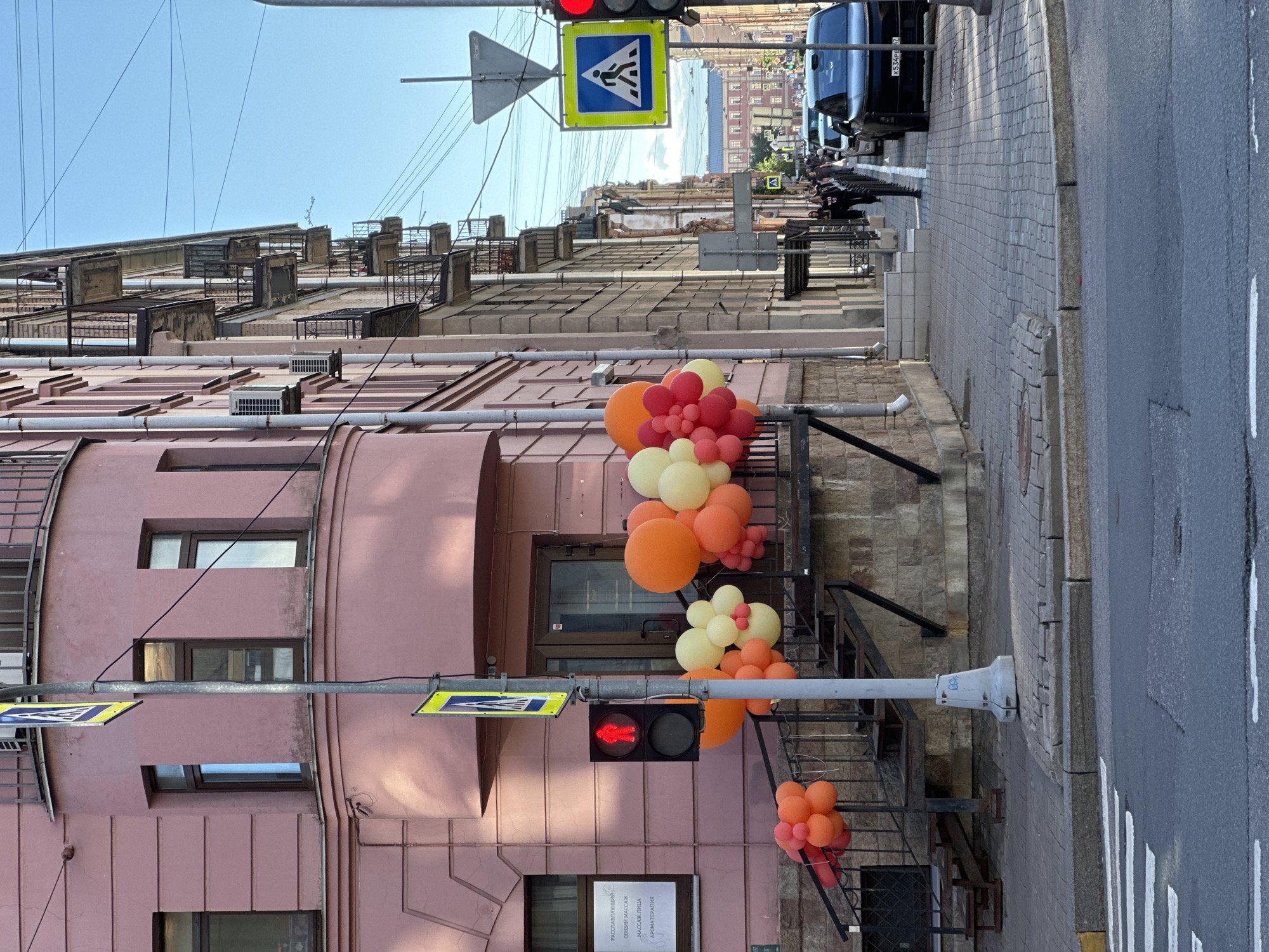 The image captures a street scene in Saint Petersburg, Russia, with a pink building on the left and a line of multi-story buildings with balconies on the right. A pedestrian crossing is visible in the foreground. A traffic light with a red pedestrian signal is illuminated. A street sign indicating a pedestrian crossing is also present. A cluster of orange and yellow balloons is attached to the railing of a stairway leading up to the pink building. Several cars are parked along the street on the right side of the image. People are walking on the sidewalk in the distance. The sky is clear and blue.
