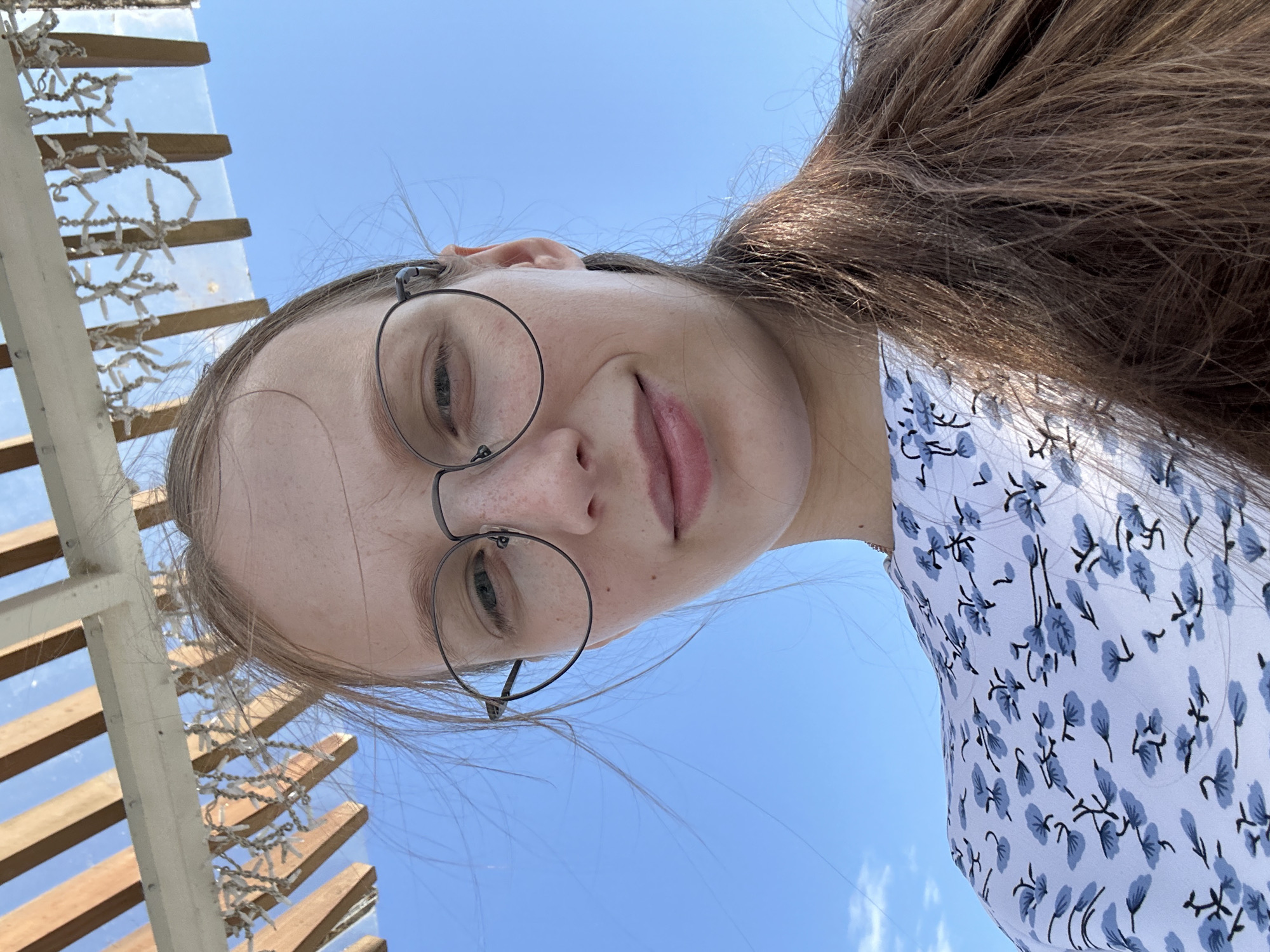 A young woman with long brown hair and glasses is looking up at the camera. She is wearing a white dress with a blue floral pattern. The background is a clear blue sky with some wooden structures and lights above her. The image appears to be taken outdoors in a sunny location, possibly a patio or pergola.