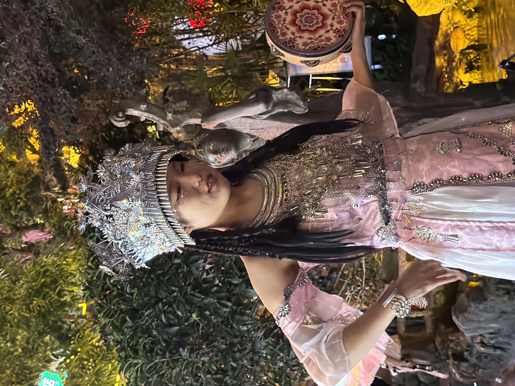 This is a photograph of a young person dressed in a traditional pink and white outfit adorned with silver embellishments. They are wearing an elaborate silver headdress and have long, dark braided hair. Their makeup includes intricate silver patterns on their face. In their right hand, they are holding a small tambourine with a decorative, floral pattern. The person is positioned in the foreground, with their body turned towards the viewer.
The background is a mix of natural and man-made elements. Lush green foliage, possibly trees and bushes, fills the lower and left portions of the image. Behind the person, there is a sculpted, greyish, possibly stone element that resembles fantastical creatures or ornate carvings. In the upper part of the image, there are glimpses of warm, colored lights, suggesting an evening or nighttime setting, possibly illuminated by artificial lighting. The overall ambiance appears festive or ceremonial. The presence of decorative lighting and elaborate costumes hints at a cultural performance or celebration.
The image appears to be taken at night or in low light conditions, as indicated by the artificial lighting and the deep shadows. The setting seems to be an outdoor or semi-outdoor space decorated for an event, possibly a garden or a themed attraction. The clothing and headdress suggest an Asian cultural context, consistent with the provided location context of Yuzhong District, China. No visible text is present in the image. The person's facial expression is somewhat neutral, with eyes closed or looking down, which could indicate focus on the performance or a serene demeanor.