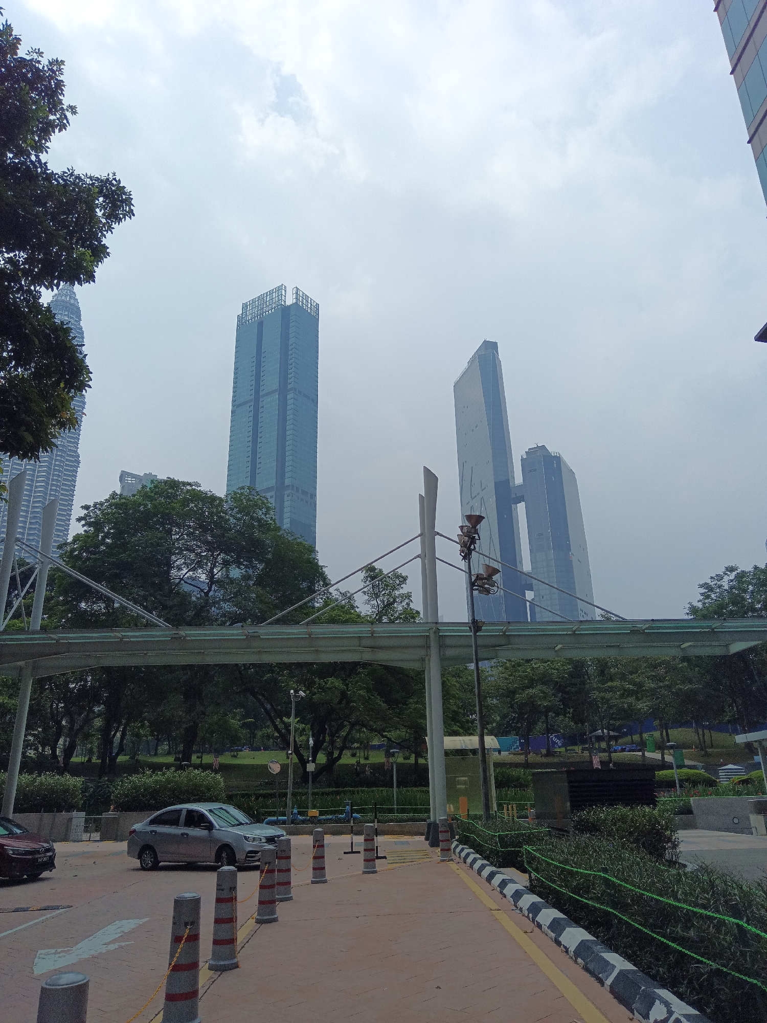 The image captures a view of the Petronas Twin Towers and other skyscrapers in Kuala Lumpur, Malaysia, on a cloudy day. In the foreground, a pedestrian walkway with bollards and a yellow line borders a road where a silver sedan is visible. A pedestrian bridge crosses over a park area with lush green trees. The background features the iconic Petronas Twin Towers, identifiable by their distinctive architecture, along with other modern high-rise buildings. The sky is overcast with light clouds, giving the scene a somewhat muted and diffused lighting.