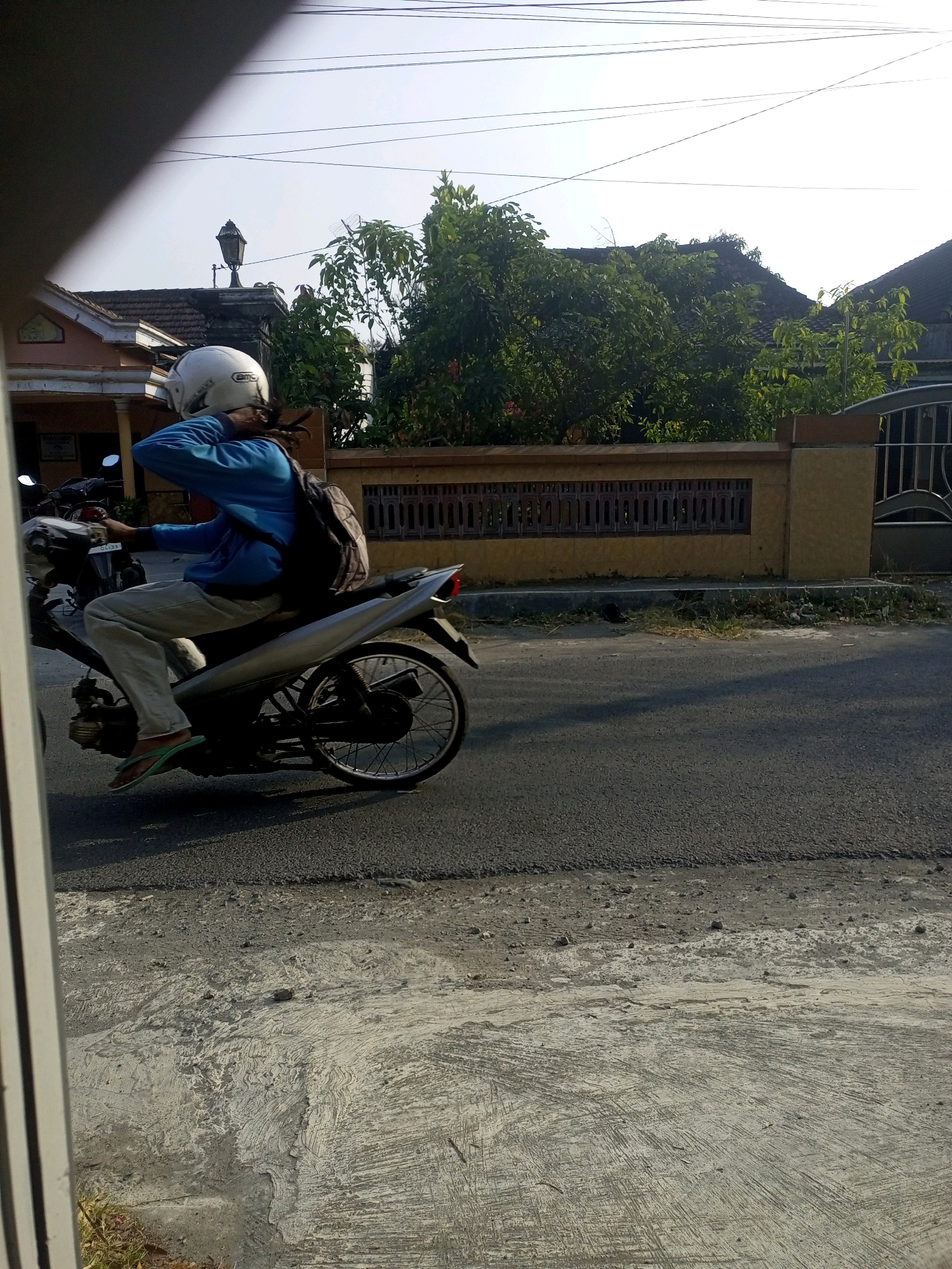 The image shows a person riding a motorcycle on a paved road in what appears to be a residential area in Ngasem, Indonesia. The person is wearing a white helmet, a blue long-sleeved shirt, light-colored pants, and green flip-flops. They have dreadlocks and are carrying a backpack. The motorcycle is silver and black.
The scene is set outdoors, with houses and lush green trees visible in the background. A decorative fence runs along the side of the road. The lighting suggests it is daytime, likely late afternoon or early morning, given the long shadows and the warm, golden light. The sky is clear and bright, indicating fair weather.
The person is in motion, their arm raised as if shielding their face or adjusting something. The overall mood is mundane, depicting a common daily activity. There is no visible text in the image, except for what might be a license plate on the motorcycle which is too blurry to read.