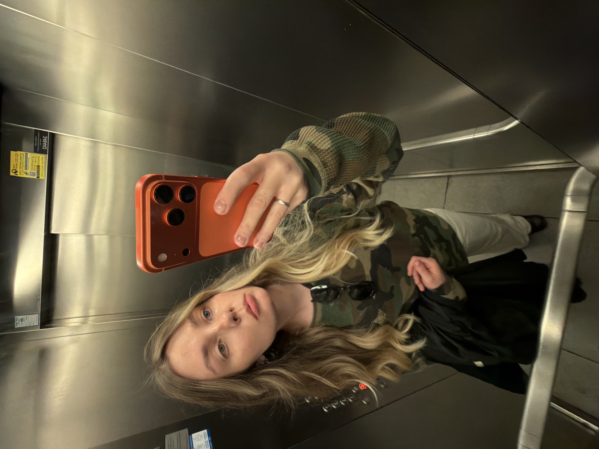 A young woman with long blonde hair is taking a selfie in an elevator. She is wearing a camouflage patterned top, white pants, and dark shoes. She is holding an orange phone with three visible lenses. Her sunglasses are on top of her shirt. The elevator has stainless steel walls and visible control buttons. Yellow sign visible on the left. It's impossible to confirm that the location is in the city of Tel-Aviv, Israel without more information.