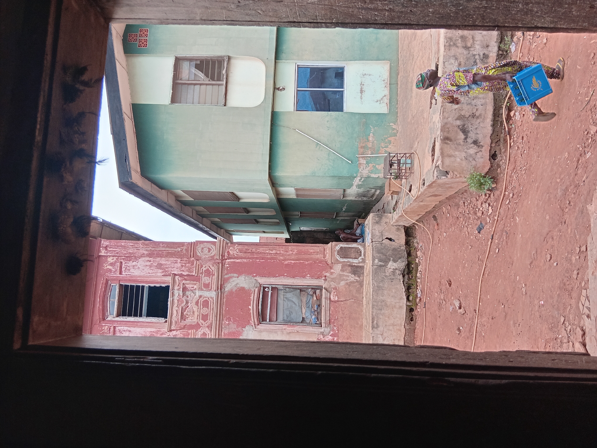 View through a wooden frame reveals a street scene in Shagamu, Nigeria. Two buildings flank the view: a green building on the top and a red building on the bottom. An adult in patterned clothing carries a blue plastic crate along a reddish-brown dirt path. A group of people sits on a step between the buildings. A yellow garden hose runs along the path.