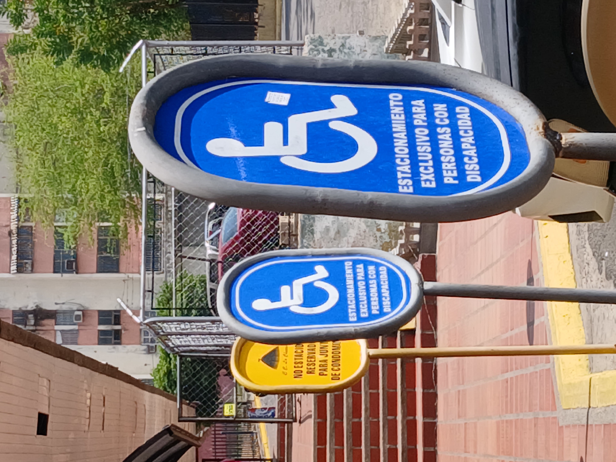 This image, captured in Guayana City, Venezuela, depicts an urban outdoor scene, likely a parking area or a street adjacent to a condominium complex during the daytime.
The main subjects are three signs mounted on poles, stacked vertically. The top two signs are identical, featuring a blue background with a white international symbol of access (a stylized person in a wheelchair). The Spanish text on these signs reads: "ESTACIONAMIENTO EXCLUSIVO PARA PERSONAS CON DISCAPACIDAD," which translates to "EXCLUSIVE PARKING FOR PEOPLE WITH DISABILITIES."
Below these blue signs is a yellow sign with black text and a downward-pointing arrow. The visible text on this sign is "NO ESTACIO RESERVAD PARA JUNT DE CONDOMIN," which is likely an abbreviation for "NO ESTACIONAR, RESERVADO PARA JUNTA DE CONDOMINIO," translating to "DO NOT PARK, RESERVED FOR CONDOMINIUM BOARD/ASSEMBLY."
In the background, there are multi-story residential or commercial buildings, some with visible windows and air conditioning units. Lush green foliage from trees is visible, as is a chain-link fence. The ground in the foreground appears to be paved with reddish bricks. The lighting is bright and clear, indicating a sunny day. No human activity or specific individuals are directly visible.