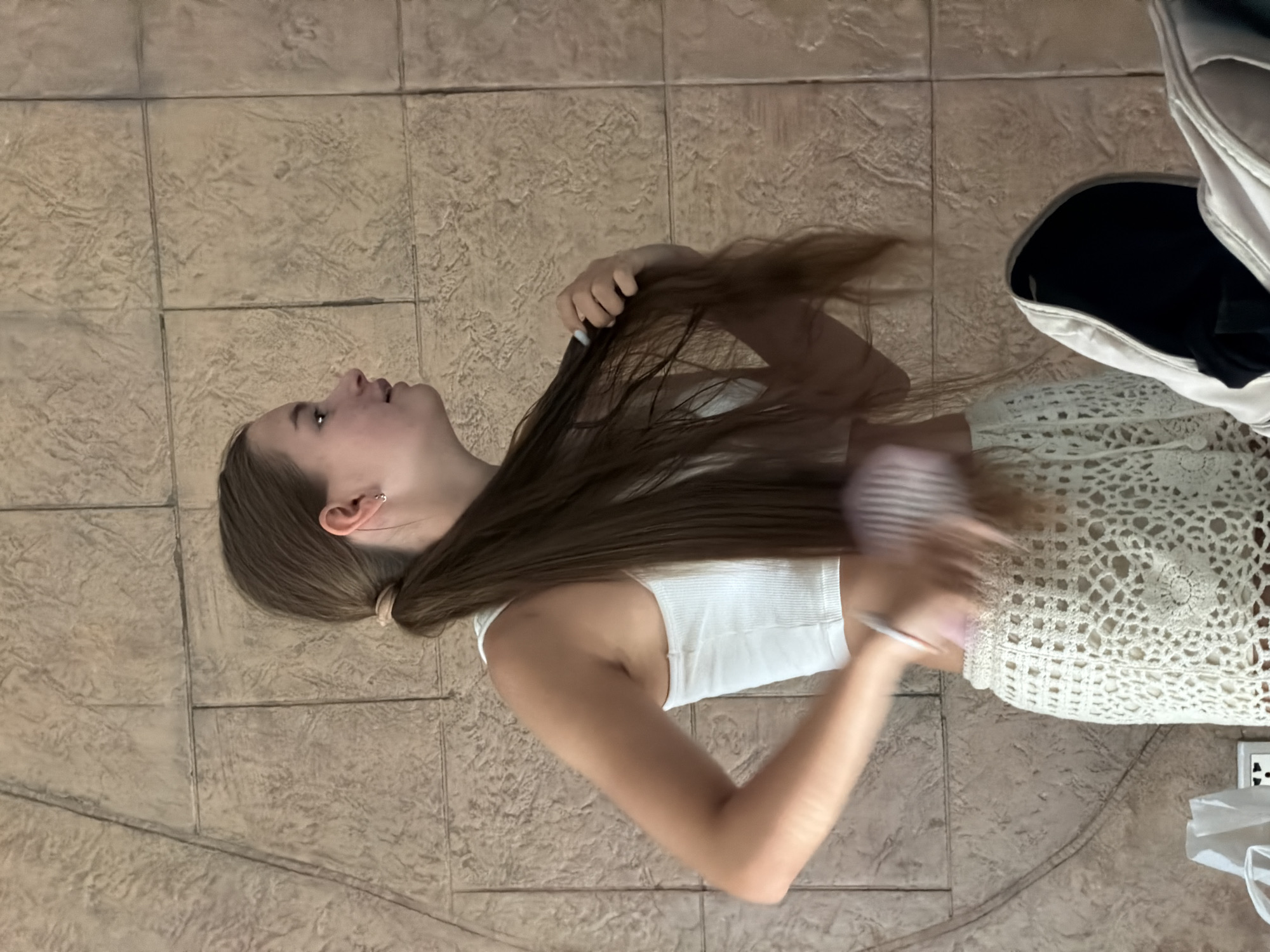 The media file shows a young person lying on a tiled floor, brushing their long, brown hair. The person is wearing a white tank top and a cream-colored crocheted skirt. Their hair is spread out around them, and they are actively brushing it. A white bag is visible to the right of the person, partially obscuring a white object with a black opening. The floor is made of large, square tiles with a textured, earthy-toned surface. The lighting suggests it is daytime. The setting appears to be indoors, possibly a home or a dressing room, with the tiled floor being a prominent feature. The person's gaze is directed upwards and to the left, and their facial expression is neutral. The overall scene is casual and domestic. The location context is Bắc Nha Trang, Vietnam.