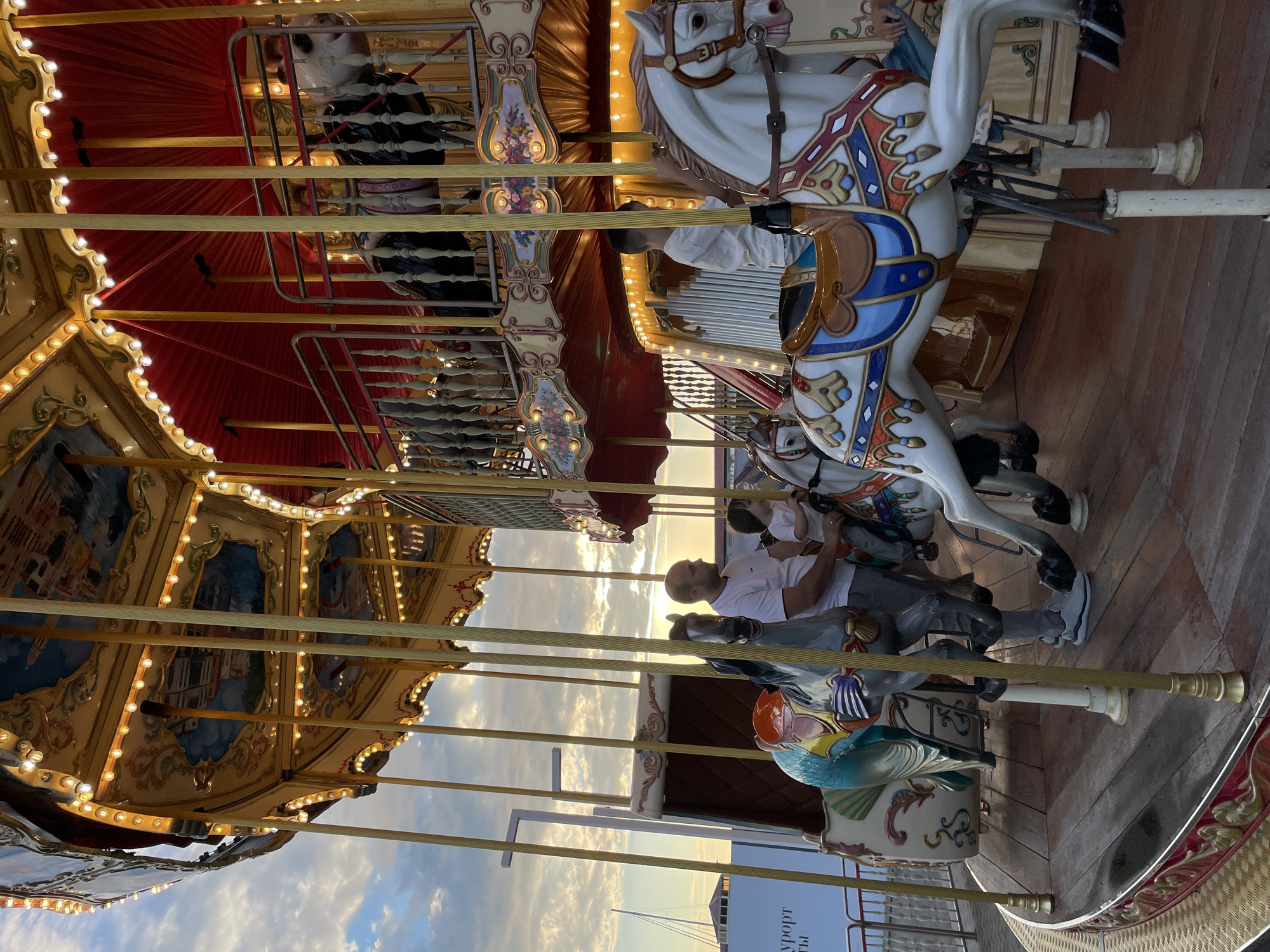 This is a photograph of a carousel, likely at a pier or waterfront location given the presence of boats in the background and the bright, cloudy sky suggesting it is daytime. The carousel is decorated with ornate carvings, gold accents, and numerous lights, indicating it is either late afternoon or early evening, with the sun beginning to set. The horses are white with colorful saddle decorations.
Several people are visible on the carousel. In the foreground, a man in a white t-shirt and grey pants is seated on one of the carousel horses. He is looking to his left, and his expression appears calm. Behind him, another person is partially visible. Further in the background, another carousel horse is visible with a person seated on it.
The carousel itself has multiple tiers, with painted panels depicting scenes that are difficult to discern clearly due to the angle and lighting. The overall mood seems peaceful and relaxed, fitting for a leisurely activity.
There is some text visible in the lower right corner of the image, on a white sign. The visible letters appear to be "УПОРТ" and "ТЫ", suggesting it might be part of a word like "Курорт" (Resort) and possibly other Cyrillic text. The location context provided is Sochi, Russia, which aligns with the possibility of this being a resort town.