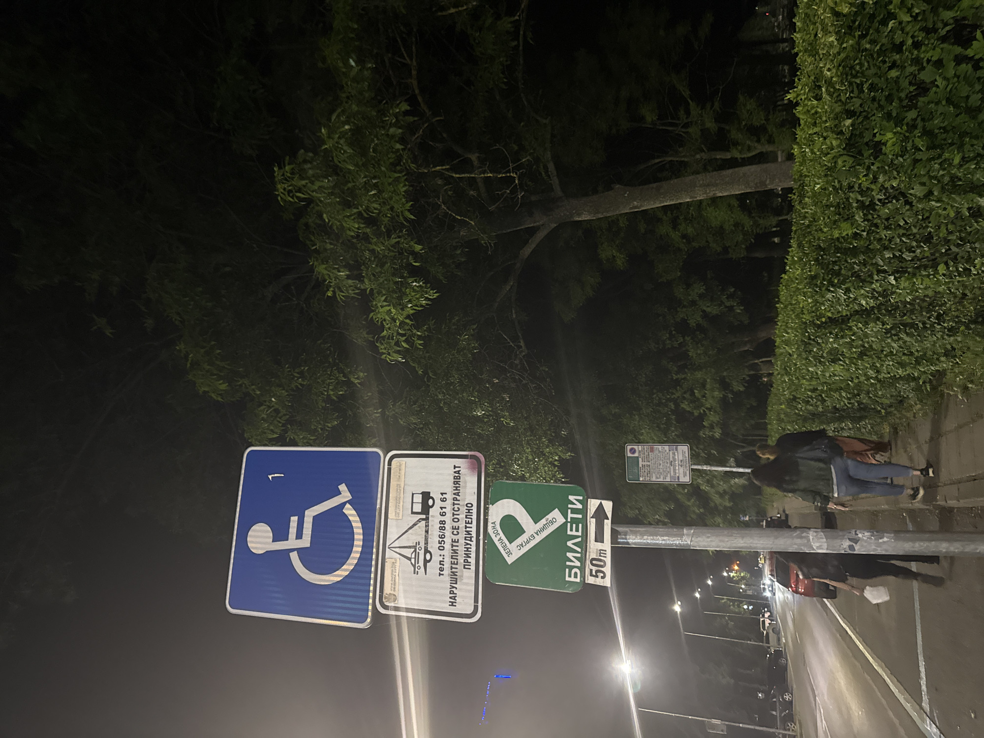 The image presents a nighttime street scene in Burgas, Bulgaria, characterized by prominent street signage and lush greenery. The perspective is slightly angled, capturing a sidewalk, a road, and dense foliage.
In the foreground, a tall street pole supports several signs. A blue sign with the international symbol for disability parking is visible on the left. Adjacent to it, a white rectangular sign features a tow truck icon, a phone number "056/88 61 61," and the Cyrillic text "НАРУШИТЕЛИТЕ СЕ ОТСТРАНЯВАТ ПРИНУДИТЕЛНО," translating to "Violators will be forcibly removed/towed." Below these, a green sign reads "ЗЕЛЕНА ЗОНА" (Green Zone) and "ОБЩИНА БУРГАС" (Burgas Municipality), indicating a controlled parking area. Further down, another green sign with an arrow points towards "БИЛЕТИ" (Tickets) at "50m" (50 meters).
Two individuals are seen walking away from the camera on the sidewalk to the right of the pole, dressed in casual clothing. The road beyond them is illuminated by streetlights and the headlights of distant cars. The entire scene is framed by dark skies and abundant green foliage, including a tall, manicured hedge bordering the sidewalk and large trees with visible trunks and branches, suggesting an urban environment with significant natural elements.