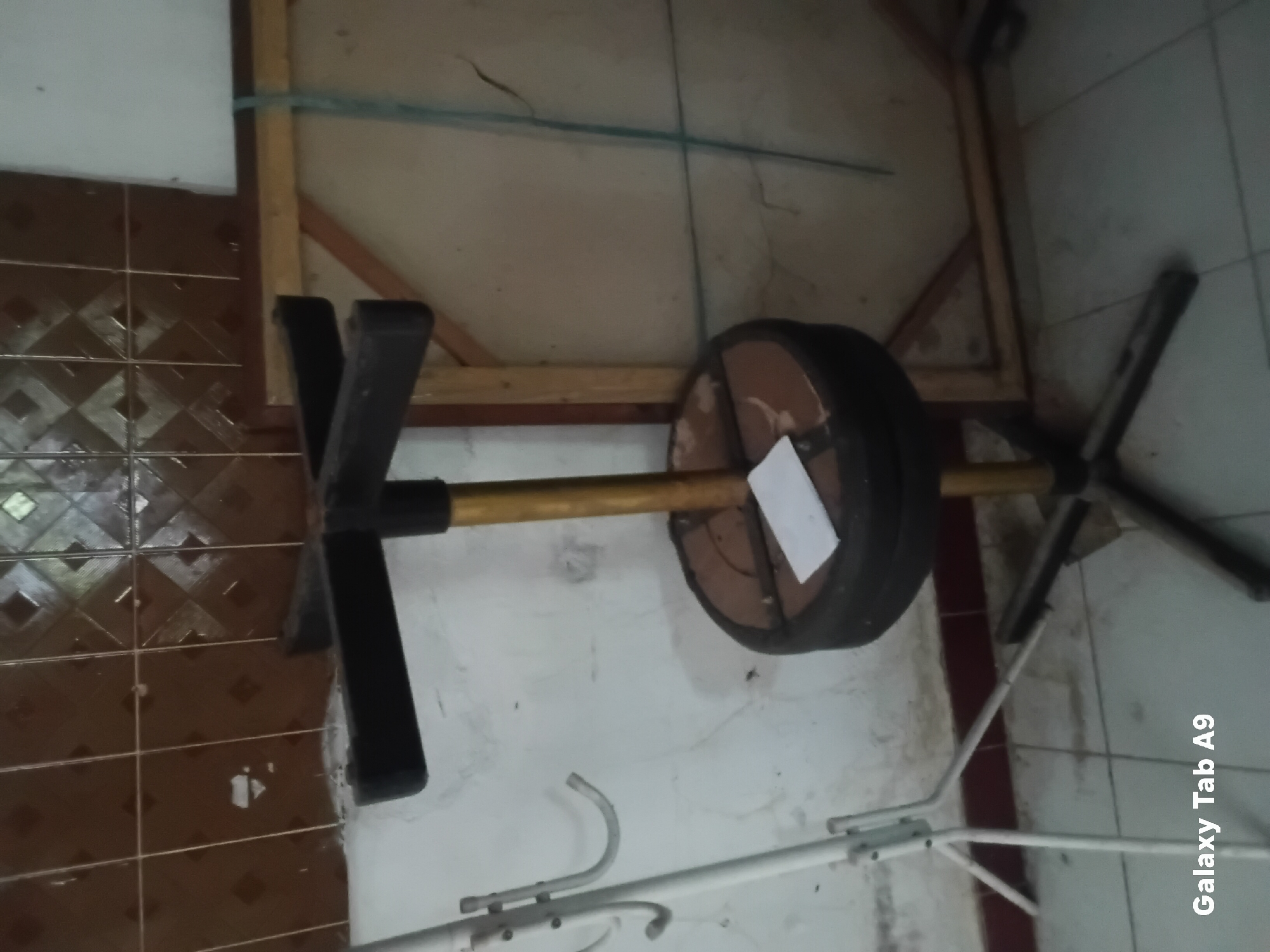 The image depicts an indoor scene, likely a room or workshop, with various objects and a tiled wall in the background. The focal point is a black and gold stool with a star-shaped base, featuring a circular padded seat. A white piece of paper rests on the seat. To the left, a white metal coat rack with curved hooks is visible. The floor is tiled, and a hint of a wooden structure or frame is present on the right. The overall impression is of a utilitarian or storage space. The location is identified as Kelutan, Indonesia, although no specific landmarks or environmental features within Kelutan are discernible from the image itself.