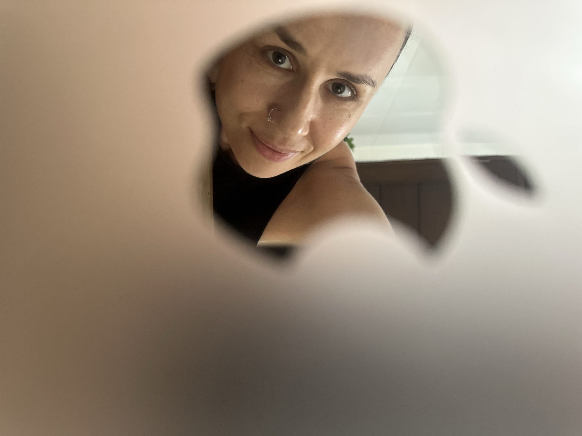 A woman is partially visible, her face looking towards the camera. She is lying down, with her head resting on her arm. Her left eye is looking directly at the camera, and she has a small smile on her face. She is wearing a black top and has a nose ring. The image is taken through a cutout in the shape of an Apple logo. The background is blurred, but it appears to be an indoor setting with some lighting and possibly furniture. The location is specified as Kuta Selatan, Indonesia.