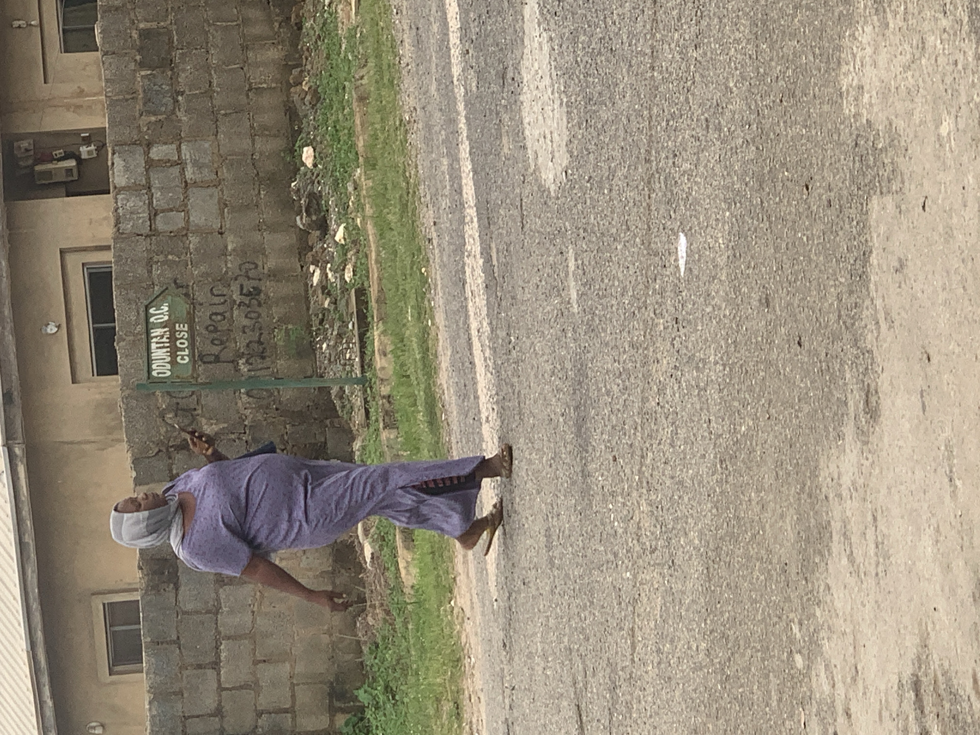 A woman walks across a paved road. She wears a light purple dress and a light gray headscarf. She is holding a mobile phone in her right hand. Behind her, a concrete block wall runs horizontally. A green sign is attached to the wall, with white text that reads "ODUNTAN O.C. CLOSE". Below the sign, "Repair" and a phone number are written on the wall in black marker. The scene is set in Abuja, Nigeria.