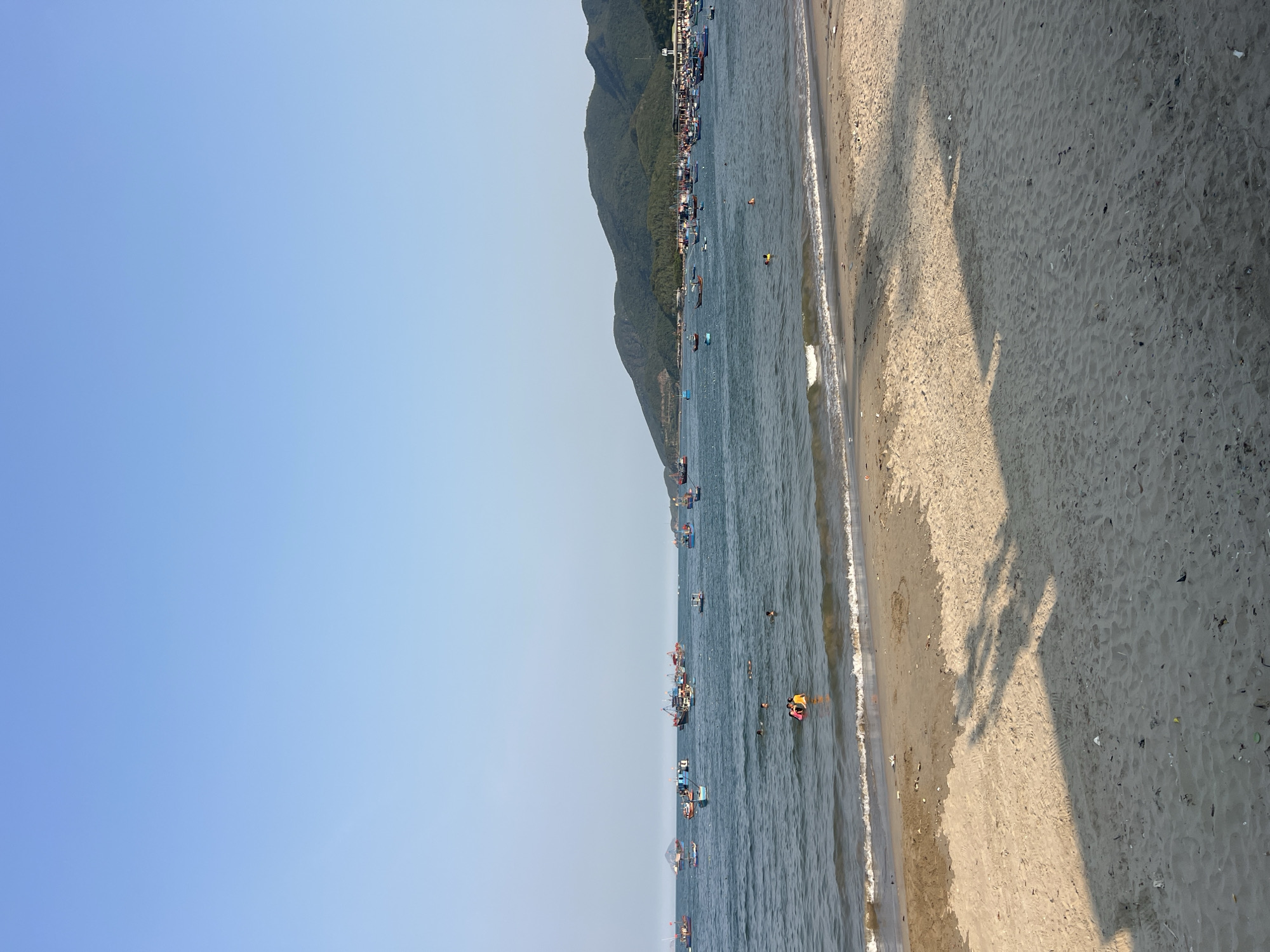 The scene is a beach in Vĩnh Lương, Vietnam. The image is taken from an elevated position, looking down on the beach. To the left, a clear blue sky fills approximately half the frame. Below the sky is the ocean, filled with boats of various sizes. Several people are in the water, some in groups. Along the shoreline, waves gently meet the sandy beach. A densely vegetated mountain range is visible in the distance. The right side of the image depicts a portion of the sandy beach, showing the shadow of people or objects, likely the photographer. The sand has some debris.
