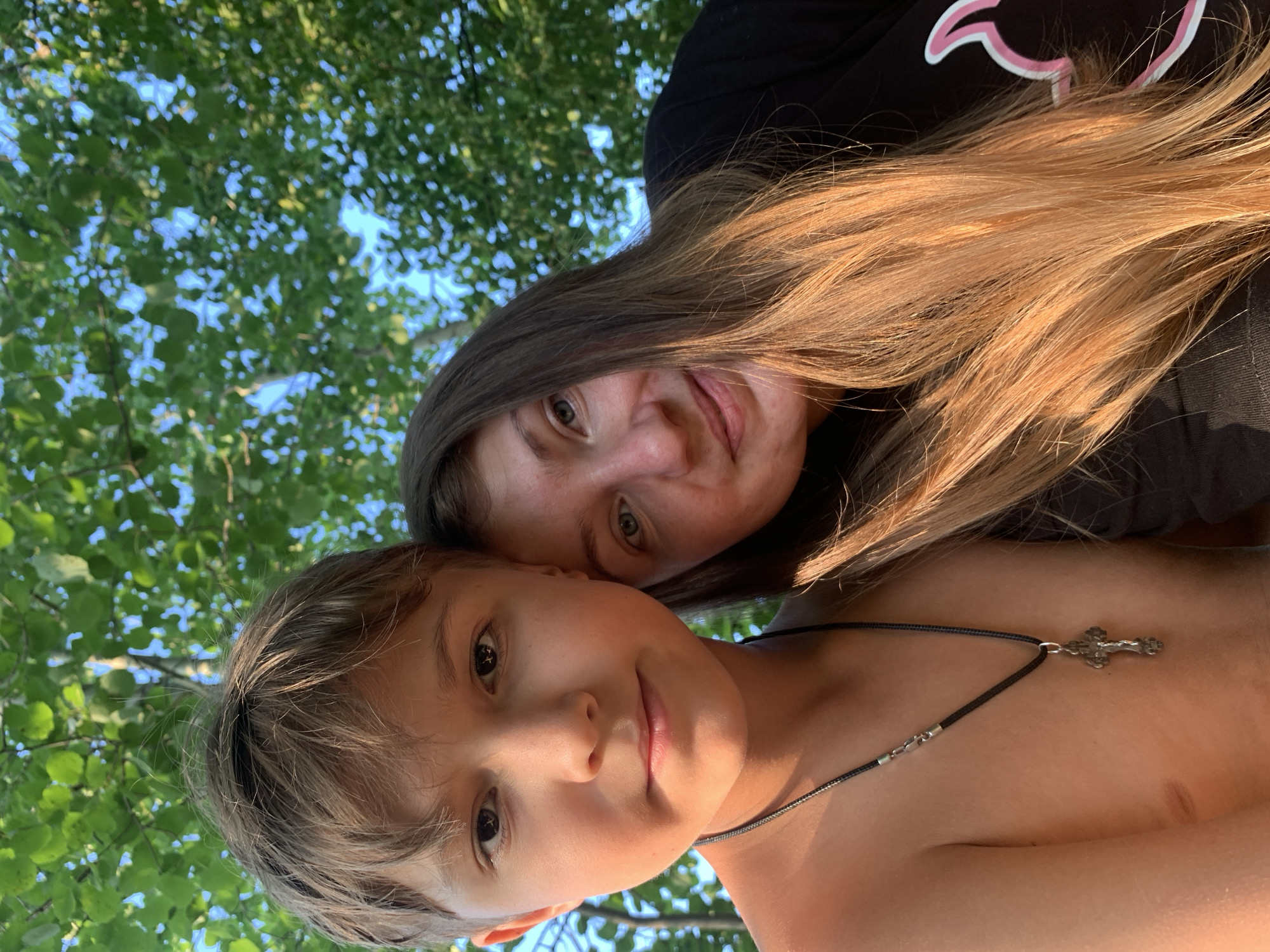 In this close-up, low-angle selfie taken outdoors in Zasechnyi, Russia, two people are pictured against a backdrop of green foliage and a bright blue sky. The image is oriented sideways.
In the foreground, a young boy is looking directly at the camera with a slight smile. His hair is a light brown, and he appears to be shirtless. He is wearing a dark cord necklace with a silver cross pendant. The sunlight highlights his face and shoulders.
Behind the boy, a woman is also looking towards the camera. Her long, light brown hair is catching the sunlight, giving it a golden hue. She is wearing a black t-shirt with a stylized pink outline of a cat on the left side of her chest. Her expression is relaxed.
The scene suggests a warm, sunny day, possibly late afternoon or early evening due to the golden light. The presence of lush green leaves indicates a location with trees, likely a park or a natural setting. The overall mood of the photograph is candid and cheerful, capturing a moment between the two individuals. No discernible text is visible in the image.