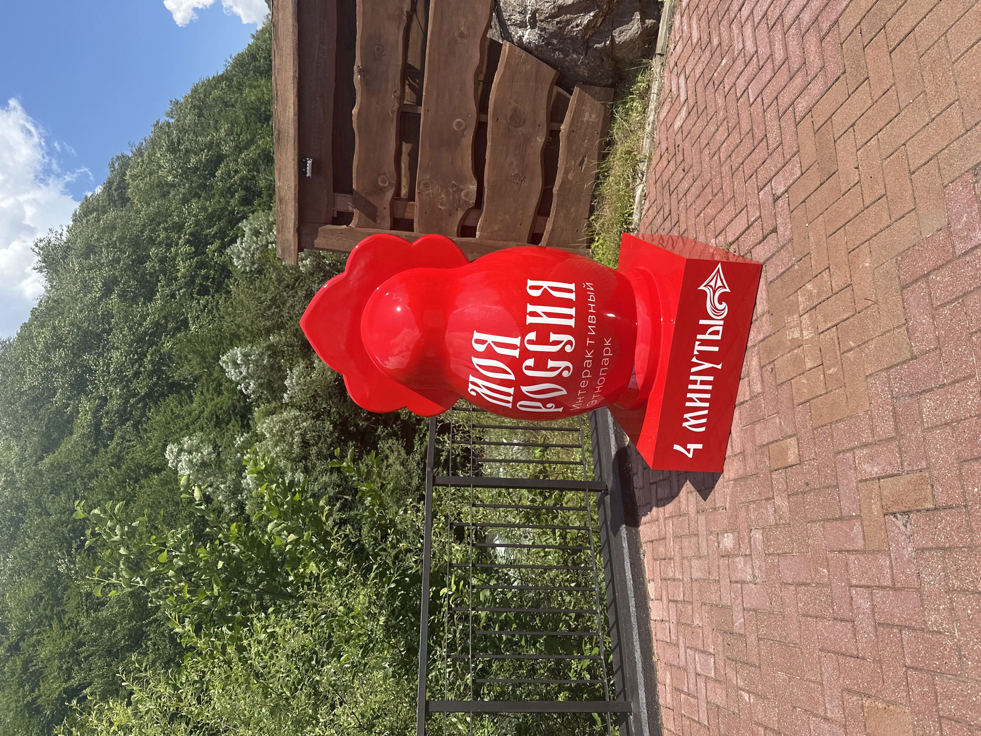 A bright red, stylized matryoshka doll sculpture stands on a paved area in Estosadok, Russia. The sculpture is positioned outdoors, with a lush green hillside and a blue sky with white clouds in the background. The doll has "МОЯ РОССИЯ" (My Russia) inscribed in white on its body, with smaller text below that reads "Интерактивный этнопарк" (Interactive Ethnopark). The base of the sculpture is also red and has "4 МИНУТЫ" (4 Minutes) written in white. The paving is made of red bricks laid in a herringbone pattern. A dark metal railing runs along the left side of the paved area, behind which is a grassy slope with more greenery. A wooden structure with horizontal planks is visible to the right of the sculpture. The sun is shining, casting shadows.