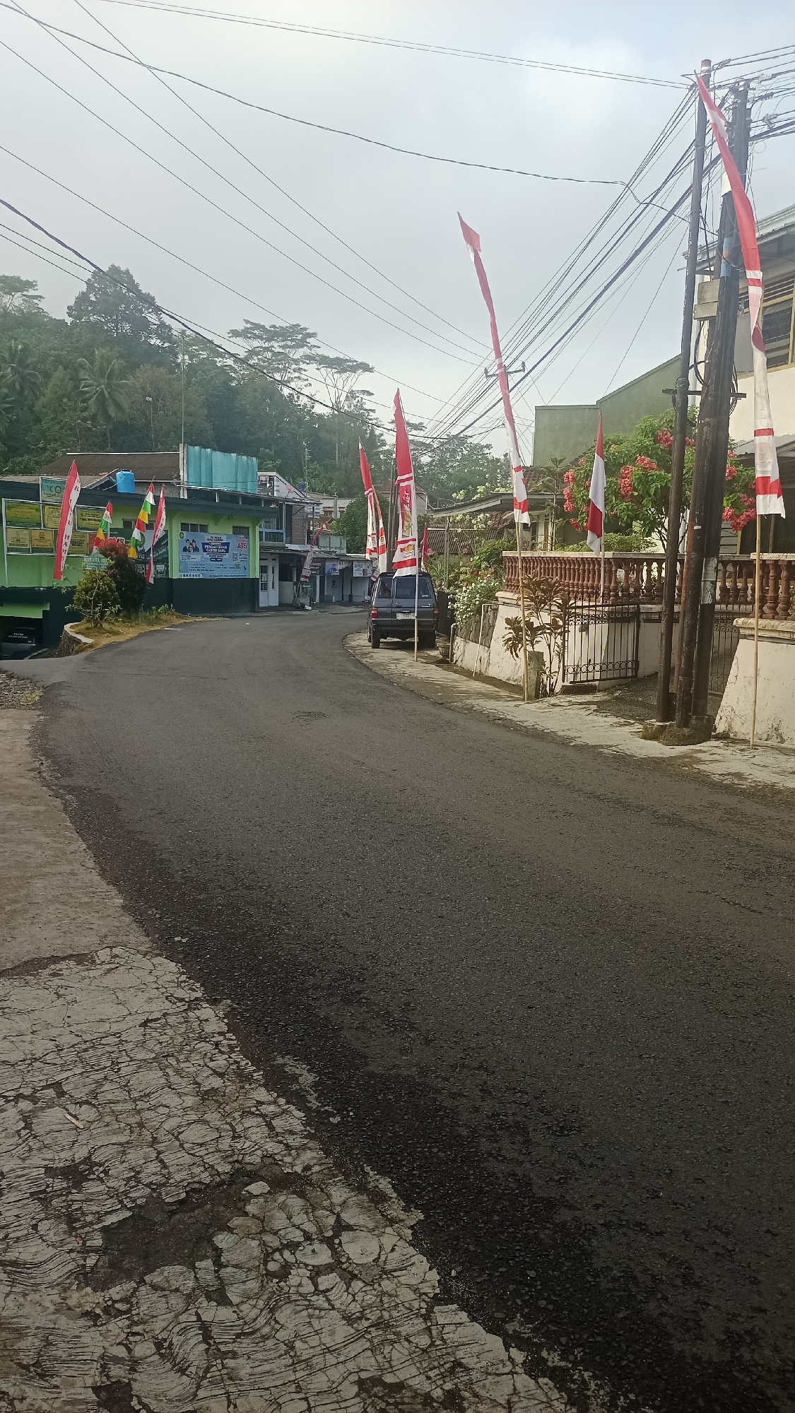 The image depicts a residential street in Karyamandala, Indonesia, under an overcast sky. The road, paved with asphalt, curves gently through a neighborhood lined with buildings and vegetation. Indonesian flags, predominantly red and white, are displayed along the roadside, attached to poles and buildings. A dark-colored SUV is parked on the right side of the street. The buildings appear to be a mix of single-story and multi-story structures, some with a green facade and others with lighter-colored walls. Dense green trees and foliage are visible in the background, suggesting a hilly or mountainous terrain. Electrical wires crisscross above the street. The concrete sidewalk on the left side of the road shows signs of age and cracking.
