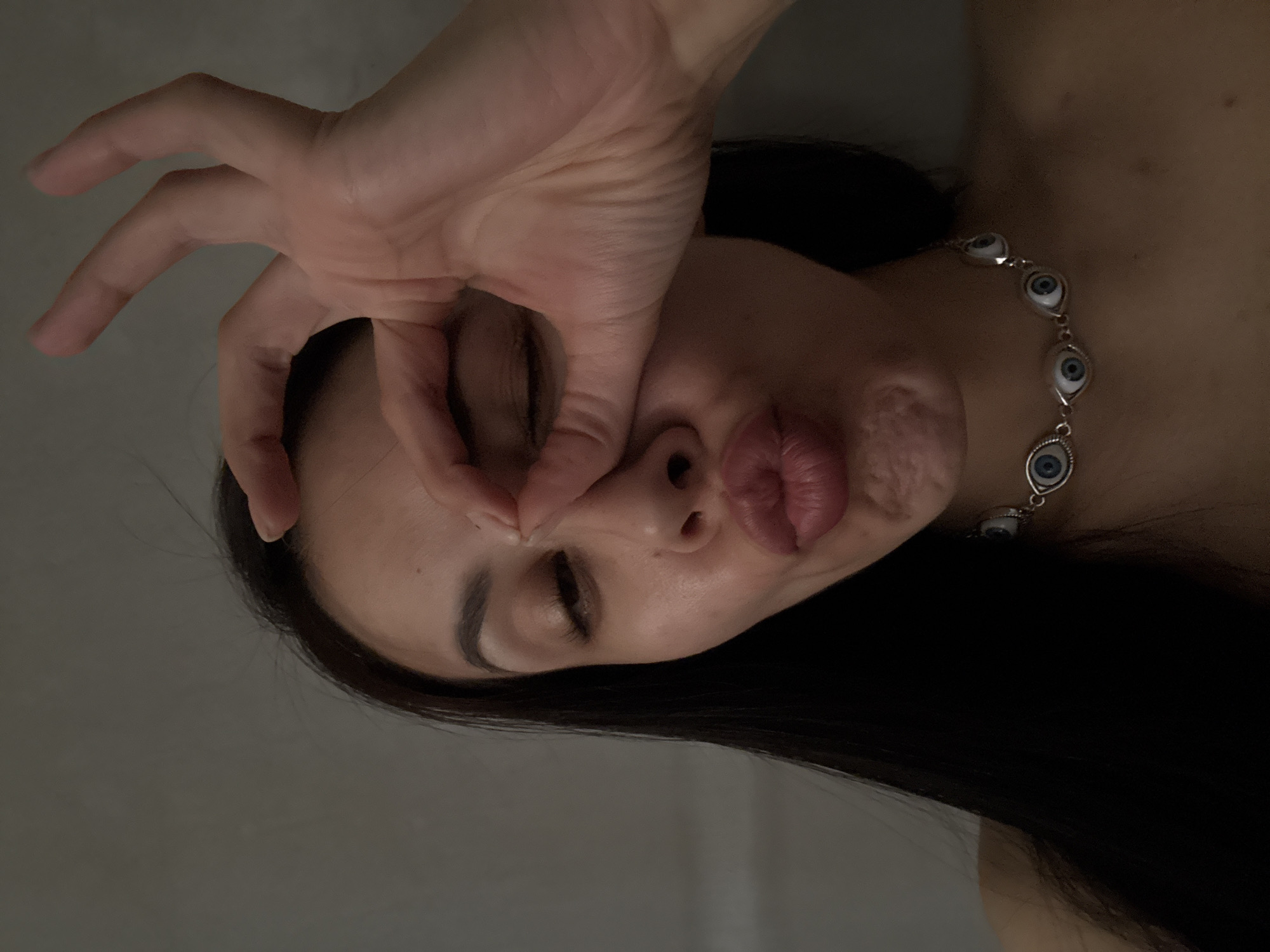 A woman is lying down, facing upward. She is positioned with her head tilted to the left. Her left hand is raised and curved, with her thumb and index finger forming a circle over her left eye. Her lips are pursed. She is wearing a necklace with eye-shaped charms. Her dark hair falls downward. The background appears to be a gray wall. The location is not discernible as Votkinsk, Russia without additional cues.
