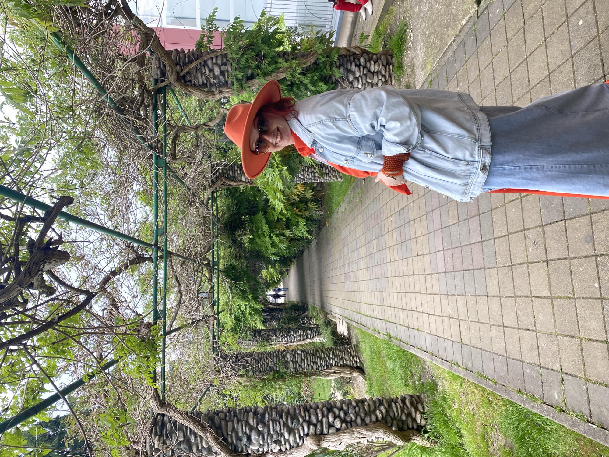 A woman stands on a paved walkway under a green metal trellis covered with vines and small branches, which form a tunnel-like structure. The walkway is lined with low walls constructed from rounded stones, with vegetation growing on both sides. The woman is wearing a light blue denim jacket, an orange hat, sunglasses, and what appears to be a bracelet made of brown beads. In the background, people can be seen walking along the walkway. The setting appears to be a park or garden in a residential area, with a building visible in the background. Given the request's mention, the location is likely in Sochi, Russia.