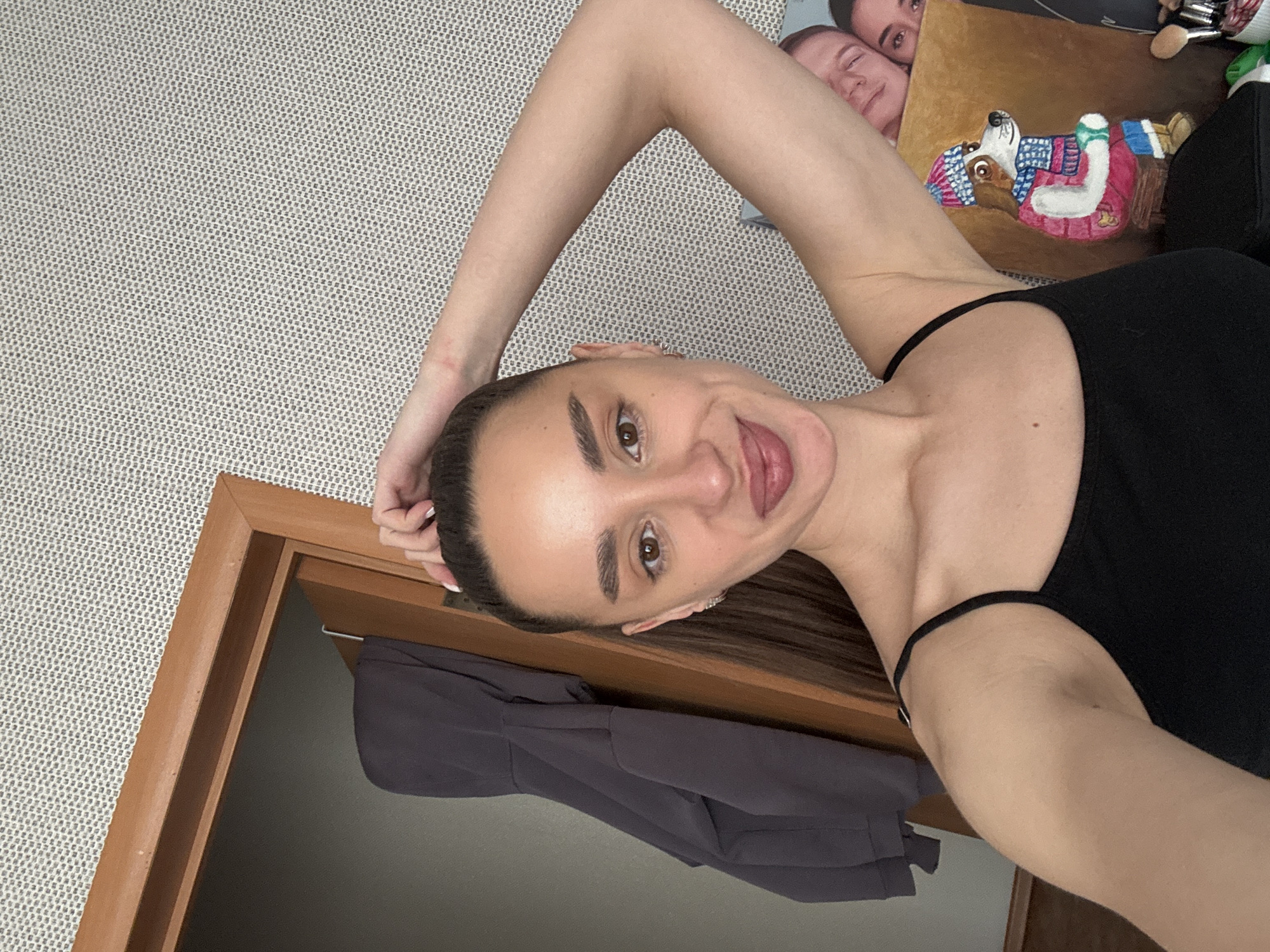 A young adult female is captured in a selfie, with the image rotated 90 degrees clockwise, suggesting she is lying down or leaning back. She holds the camera overhead, looking directly at it with a playful, open-mouthed expression, revealing her tongue. Her dark hair is pulled back, and she wears a simple black tank top with thin straps.
The background to her left features a light gray, textured wall. Above her right shoulder, various personal items are visible on a surface. A prominent painting depicts a dog wearing a blue hat, a striped scarf, and a red coat. Behind this, a portion of another painting or print shows two faces. To the right of the dog painting, several makeup brushes or similar small tools are arranged.
In the bottom left corner, a wooden door frame opens into a darker room where a dark gray garment, possibly a jacket or hoodie, hangs on a hook. The setting appears to be an indoor domestic space, likely a bedroom or living area. The woman's expression conveys a cheerful and relaxed demeanor. No visible text is present in the image.