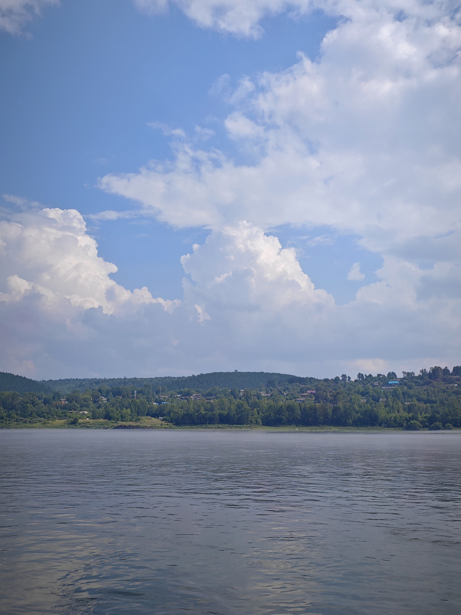 The image shows a wide river with gentle ripples, reflecting the sky. A thin layer of mist hovers over the water's surface near the shore. The opposite bank is lined with a dense green forest that ascends a hilly landscape. Dotted throughout the trees, and visible on the slopes, are houses, indicating a settlement. Above the treeline, the sky is a bright blue, populated by large, fluffy white clouds. The overall scene depicts a tranquil, natural environment with signs of human habitation on the distant shore, suggesting a location in or near a rural area like Pristan', Russia.