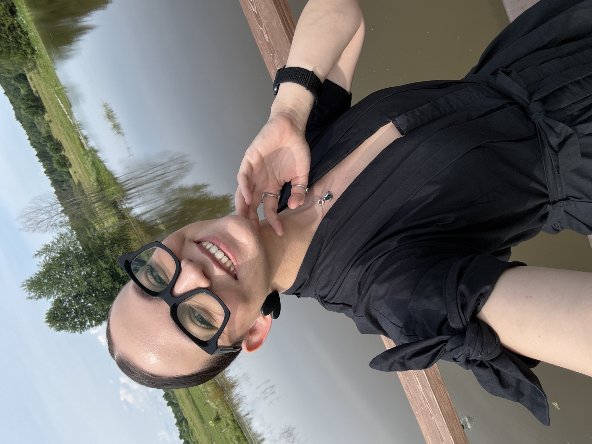 A woman with dark hair pulled back and wearing large black glasses takes a selfie outdoors. She has on a black dress with a deep neckline and a bow tie at the waist. She is also wearing a necklace with a blue pendant and a black watch. Her left hand is resting on her chin. Behind her is a body of water with trees and a grassy field in the background. The setting appears to be near the city of Svetloye, Russia.