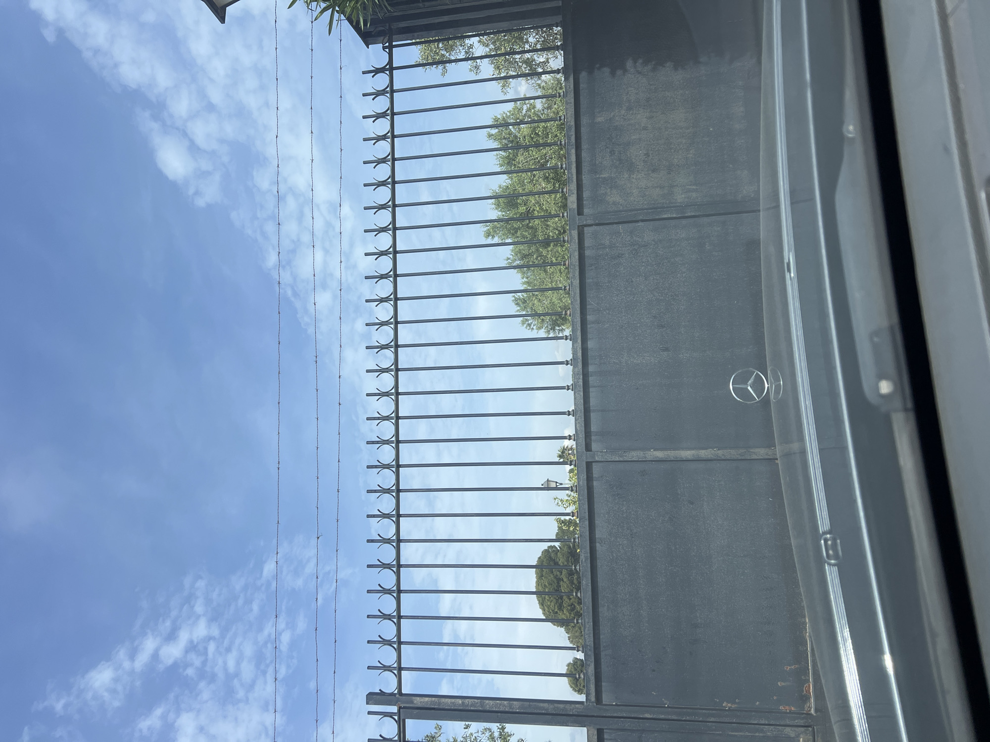 The image captures an outdoor scene, seemingly taken from inside a vehicle, looking out through a window at a tall, dark gate and the sky beyond. The perspective suggests a slightly upward gaze.
In the foreground, a distinct reflection of a Mercedes-Benz steering wheel logo is visible in the lower right portion of the image, superimposed on the gate. This indicates the photograph was taken from within a car. On the far right, parts of the car's interior, possibly a window frame or door panel, are visible.
The main subject is a substantial dark metal gate or fence. The lower section of the gate appears solid. Above this, there's a panel composed of vertical metal bars, each featuring a decorative, C-shaped or semi-circular element at its top. Further above this section, several strands of barbed wire are stretched horizontally, acting as an additional security feature.
In the background, a bright blue sky dominates the upper part of the frame, adorned with scattered white, wispy clouds. Through the openings in the gate's railings and above its structure, numerous green trees with dense foliage are visible. Some leafy vegetation also peeks into the frame from the top left corner, possibly belonging to a nearby building or overgrown plants.
The scene is brightly lit, indicating it is daytime with sunny weather. There is no human activity depicted, contributing to a static and calm atmosphere. The image provides a glimpse of a secured property boundary in Vila-seca, Spain.