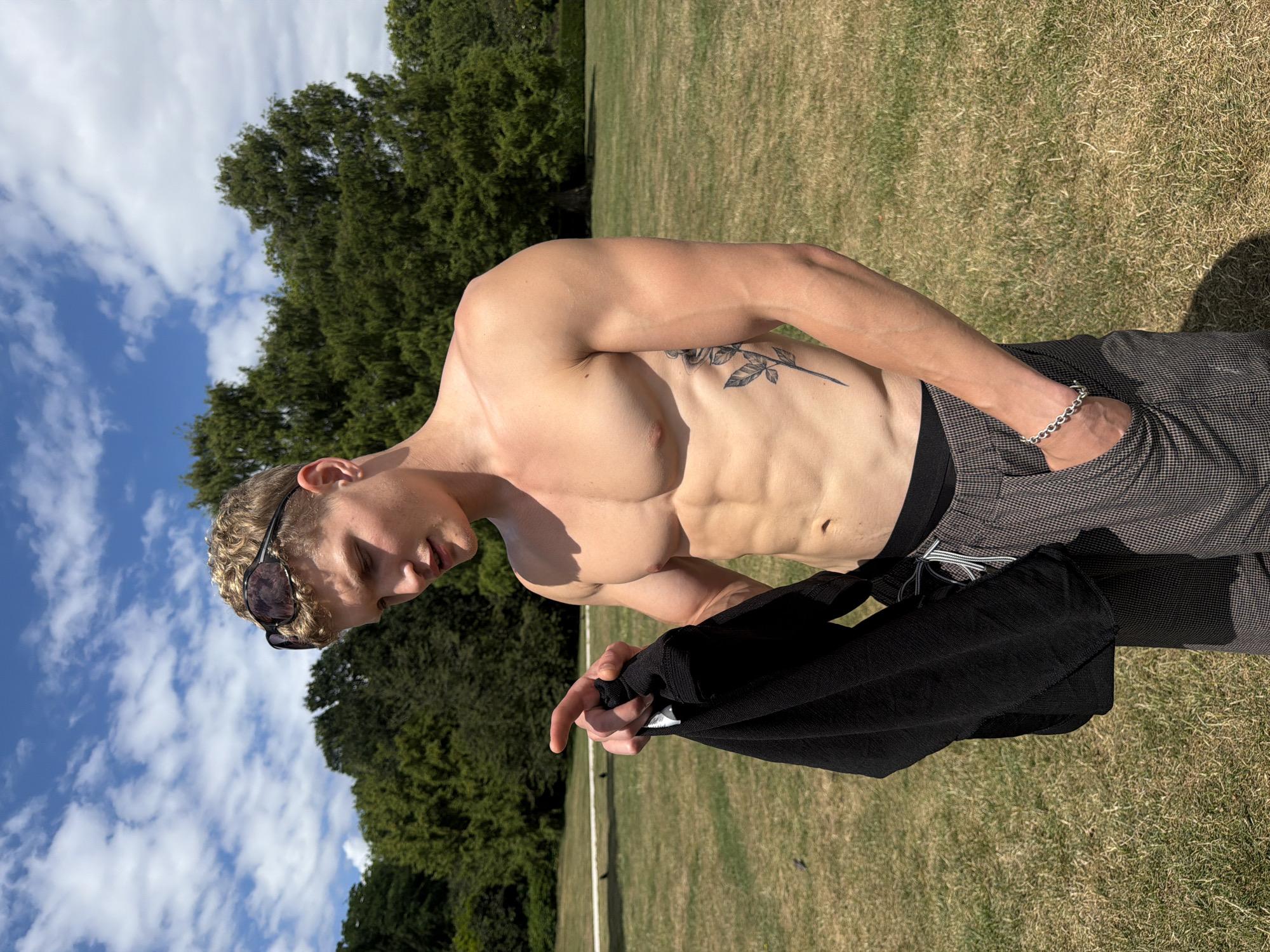 A young Caucasian male with blonde curly hair and sunglasses on his head is standing in a grassy park on a sunny day. He is shirtless, displaying a toned physique and a rose tattoo on his left side. He is holding a black garment in his left hand and pointing with his right index finger. He is wearing grey patterned pants with a black waistband and a silver chain bracelet on his left wrist. The background consists of lush green trees and a blue sky with white clouds. There are two birds on the grass in the mid-ground. The location is likely a public park in the city of London, United Kingdom, given the presence of manicured lawns and mature trees, characteristic of London's many green spaces.