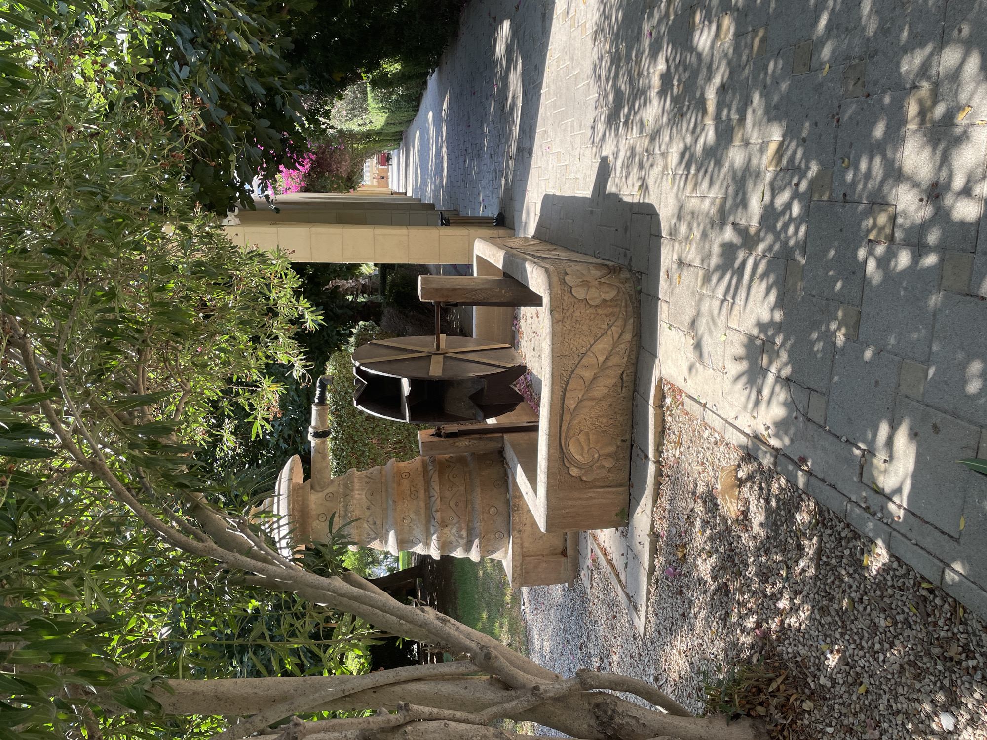 A stone water feature with a decorative wheel and trough is situated next to a paved walkway lined with trees and bushes. The water feature is made of light-colored stone and has intricate carvings. The walkway is paved with rectangular stones and is shaded by the surrounding trees, casting dappled shadows on the ground. In the background, there is a row of arched structures with stone pillars, suggesting an architectural element of the location. The environment is outdoors, with lush greenery and a clear sky, indicating a sunny day. The scene is serene and tranquil, with no people or animals visible. The presence of a water feature and decorative elements suggests it might be a garden, park, or courtyard. The overall impression is one of a well-maintained and aesthetically pleasing outdoor space.