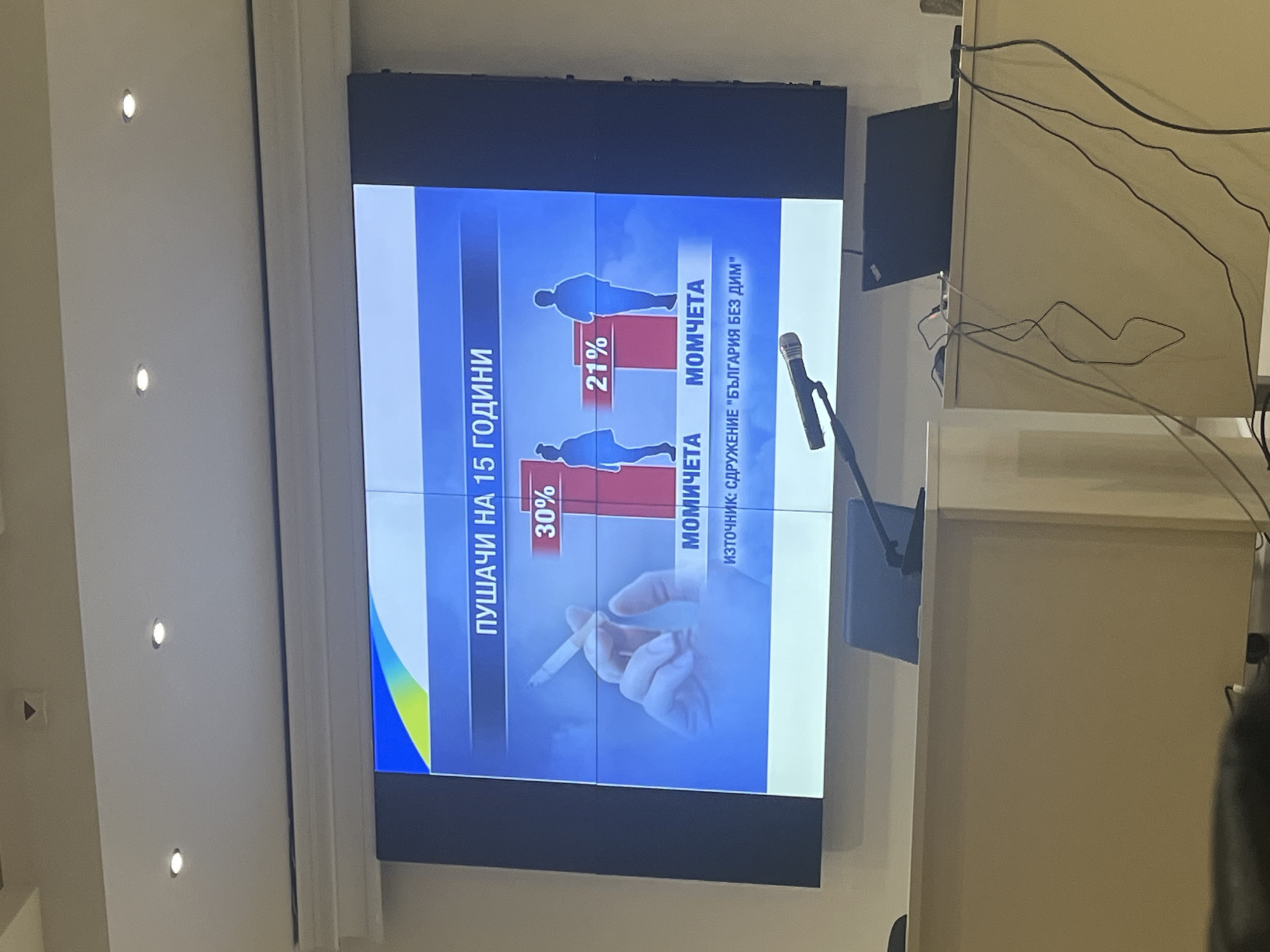 The image displays a large video wall or multiple interconnected screens, vertically mounted on a light-colored wall, likely in a conference room or a presentation hall in Plovdiv, Bulgaria. The screen content, which is oriented sideways in the photograph, depicts statistics about smoking.
The presentation slide is titled "ПУШАЧИ НА 15 ГОДИНИ" (SMOKERS AT 15 YEARS OLD). Below this title, a bar graph indicates that 30% of "МОМИЧЕТА" (GIRLS) and 21% of "МОМЧЕТА" (BOYS) are smokers at this age, accompanied by gender-specific silhouettes. The lower part of the screen features a close-up image of a hand holding a lit cigarette. The source of the information is attributed to "ИЗТОЧНИК: СДРУЖЕНИЕ 'БЪЛГАРИЯ БЕЗ ДИМ'" (SOURCE: ASSOCIATION "BULGARIA WITHOUT SMOKE"). The screen's background is primarily blue, with a colorful gradient in the bottom left corner.
To the left of the screen, several recessed spotlights are visible on the wall. To the right, a microphone on a stand suggests a speaker is addressing an audience. Behind the microphone, a smaller black monitor and a cluster of wires are visible against a light-colored surface. The setting implies a formal event or public awareness campaign concerning youth smoking, possibly a press conference or seminar. The time of day is not discernible as the scene is indoors with artificial lighting.