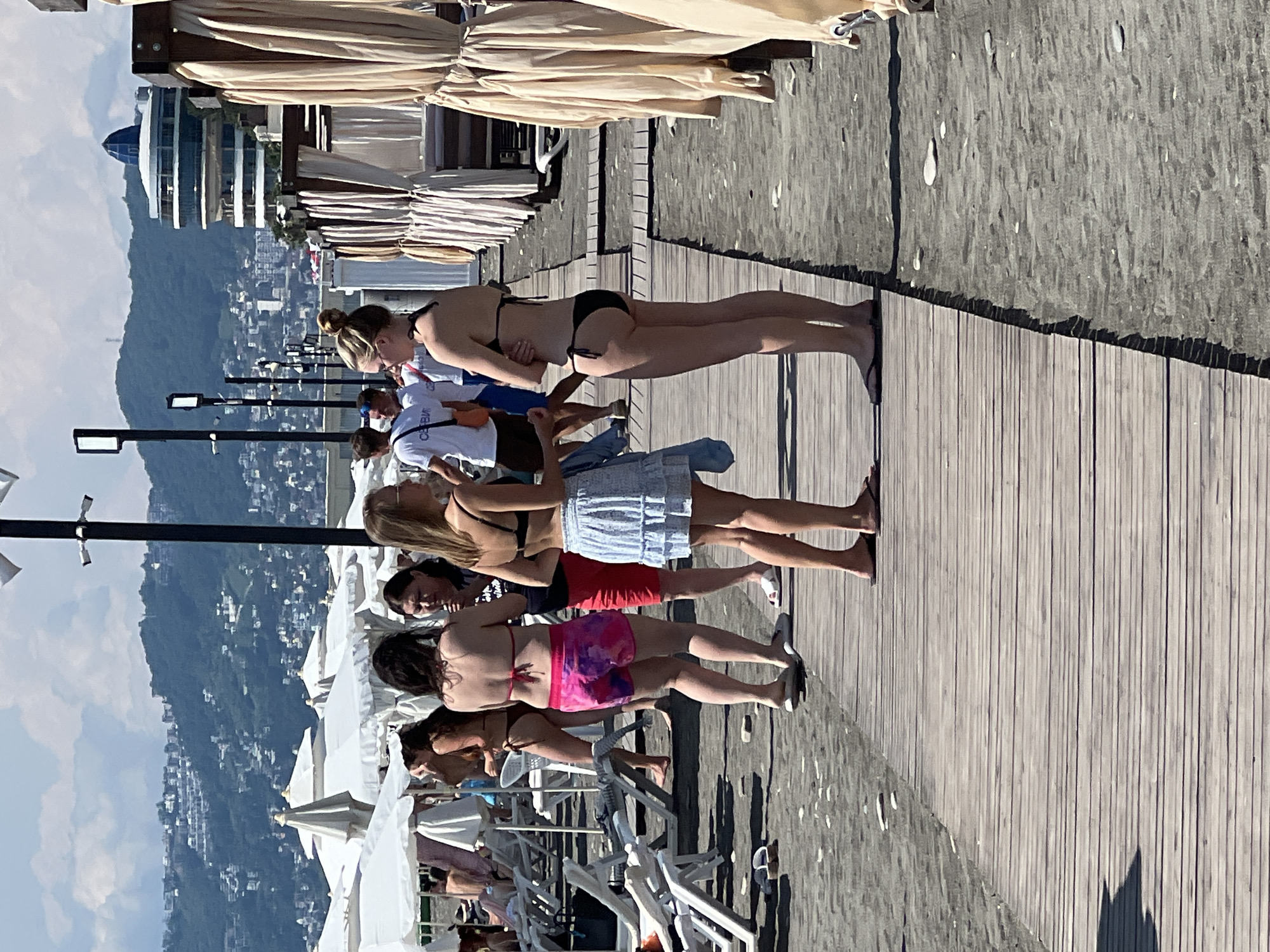 The image depicts a sunny day at a beach in Sochi, Russia. A wooden boardwalk runs diagonally across the foreground, separating the dark sand from lounge chairs and white umbrellas further back. Several people are visible on the boardwalk and near the beach.
In the mid-ground, three women are conversing. The woman on the left is wearing a brown bikini. The woman in the center is wearing a red and purple patterned skirt over a bikini bottom and a red bikini top. The woman on the right is wearing a light blue skirt over a bikini bottom and a black bikini top. To the right of these women, another woman in a black bikini is standing.
In the background, there are more beachgoers, lounge chairs, and umbrellas. Beyond the beach, a hillside covered with buildings and a distinctive modern circular building with a blue dome are visible, characteristic of Sochi's cityscape. The sky is bright blue with some scattered white clouds. The overall scene is of a lively beach environment.