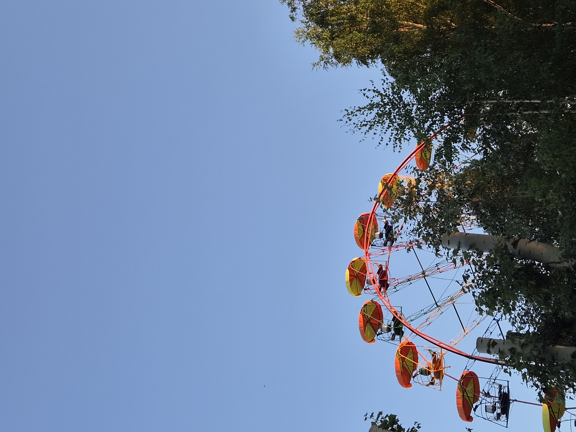The image captures a segment of a Ferris wheel, its red and yellow gondolas outlined against a clear blue sky. Trees partially obscure the lower portion of the ride, with the tops of their green foliage visible. The Ferris wheel appears to be in a park or amusement area. No people are clearly discernible within the gondolas or the surrounding area. The overall scene suggests an outdoor setting during daylight hours. This Ferris wheel is located in the city of Chaikovsky, Russia.