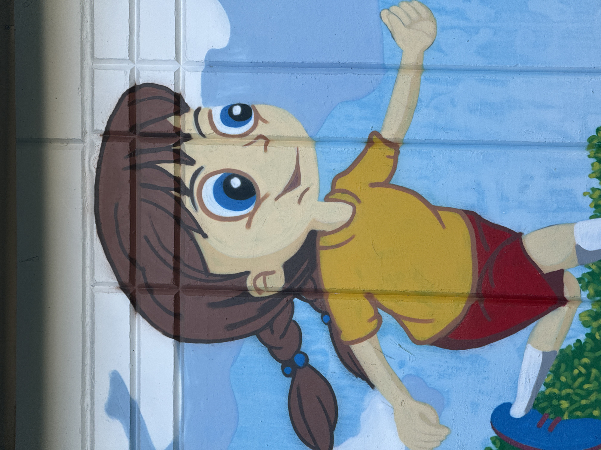 This is a close-up, medium shot, eye-level, outdoor photograph of a mural painted on a brick wall. The mural depicts a cartoon girl with brown braided hair, blue eyes, and a yellow t-shirt with red shorts. She is shown in profile, running with her arms outstretched, against a blue sky with white clouds. Green foliage is visible at the bottom of the frame. The mural is painted in a simple, colorful style, with bold outlines and flat areas of color. The brick wall behind the mural has visible lines and texture. The lighting is bright and even, suggesting daylight. There are no people or animals present in the photograph, only the painted image.