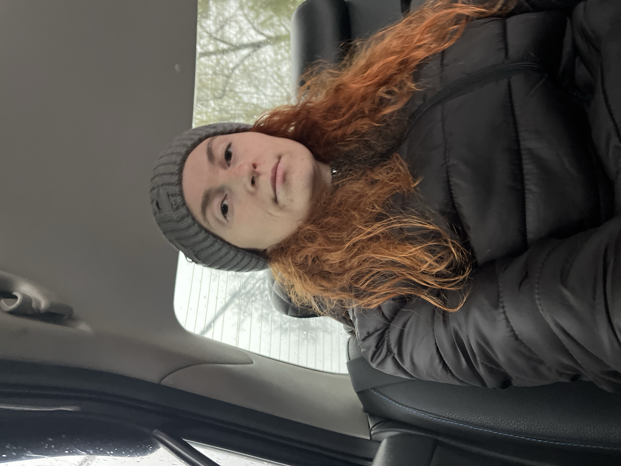 A medium shot of a young adult with red curly hair wearing a grey beanie and a black puffer jacket inside a vehicle. Rain streaks are visible on the car windows. The location is likely in Chaikovsky, Russia, but this cannot be definitively confirmed from the image.
