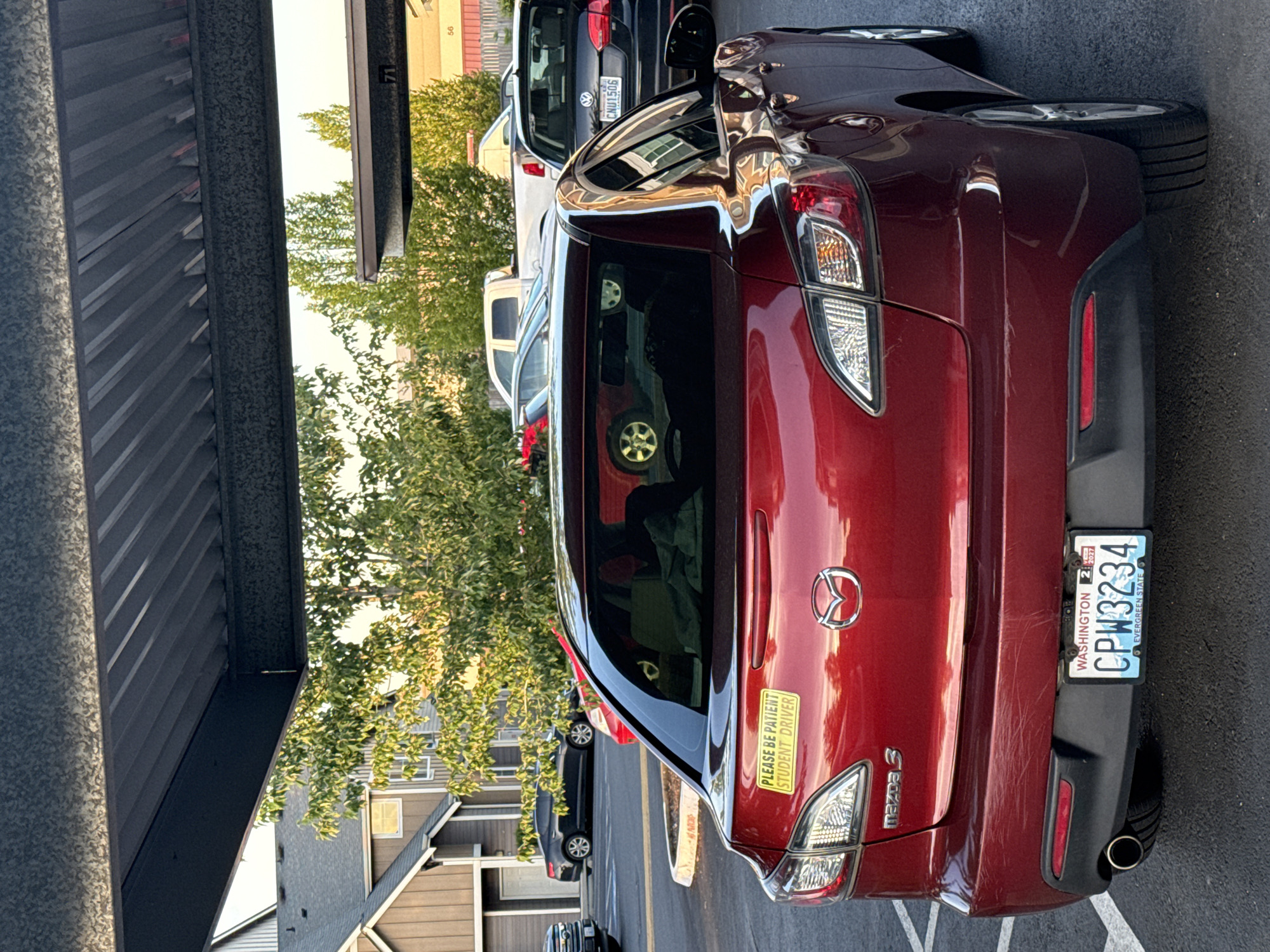 The image captures the rear of a red Mazda 3 parked under a covered structure, likely in a residential or commercial parking lot. The car is facing away from the viewer, and its license plate reads "CPW3234" from Washington state. A yellow sticker on the rear windshield says "PLEASE BE PATIENT STUDENT DRIVER." In the background, other vehicles, trees, and a multi-story building are visible. The overall environment suggests an outdoor setting in daylight.