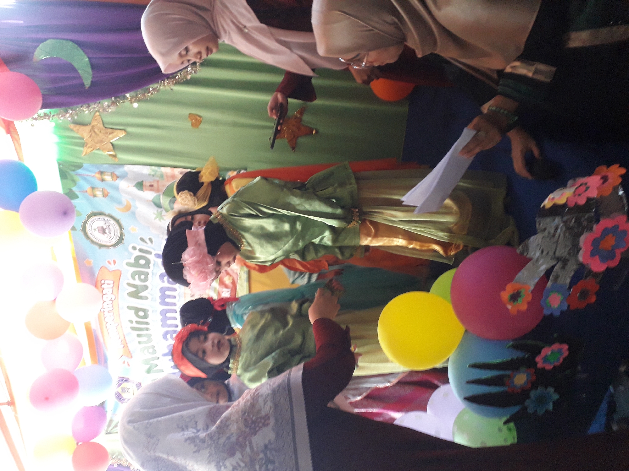 The image captures an indoor event, possibly a school or community gathering, in Cankuang, Indonesia, indicated by the banner that reads "Peringati Maulid Nabi Muhammad." The scene is decorated with balloons of various colors, including pink, yellow, blue, and white, and a green curtain serves as a backdrop. Several children, dressed in traditional attire, are positioned in the center of the frame. Some are wearing hijabs and long dresses in green and yellow. Two adult women are also present. One woman, on the right, is wearing a light pink hijab and holding a mobile phone. Another woman, on the left, is wearing a patterned hijab and a maroon top, with her arms extended, holding a piece of paper. The overall atmosphere suggests a celebratory event.