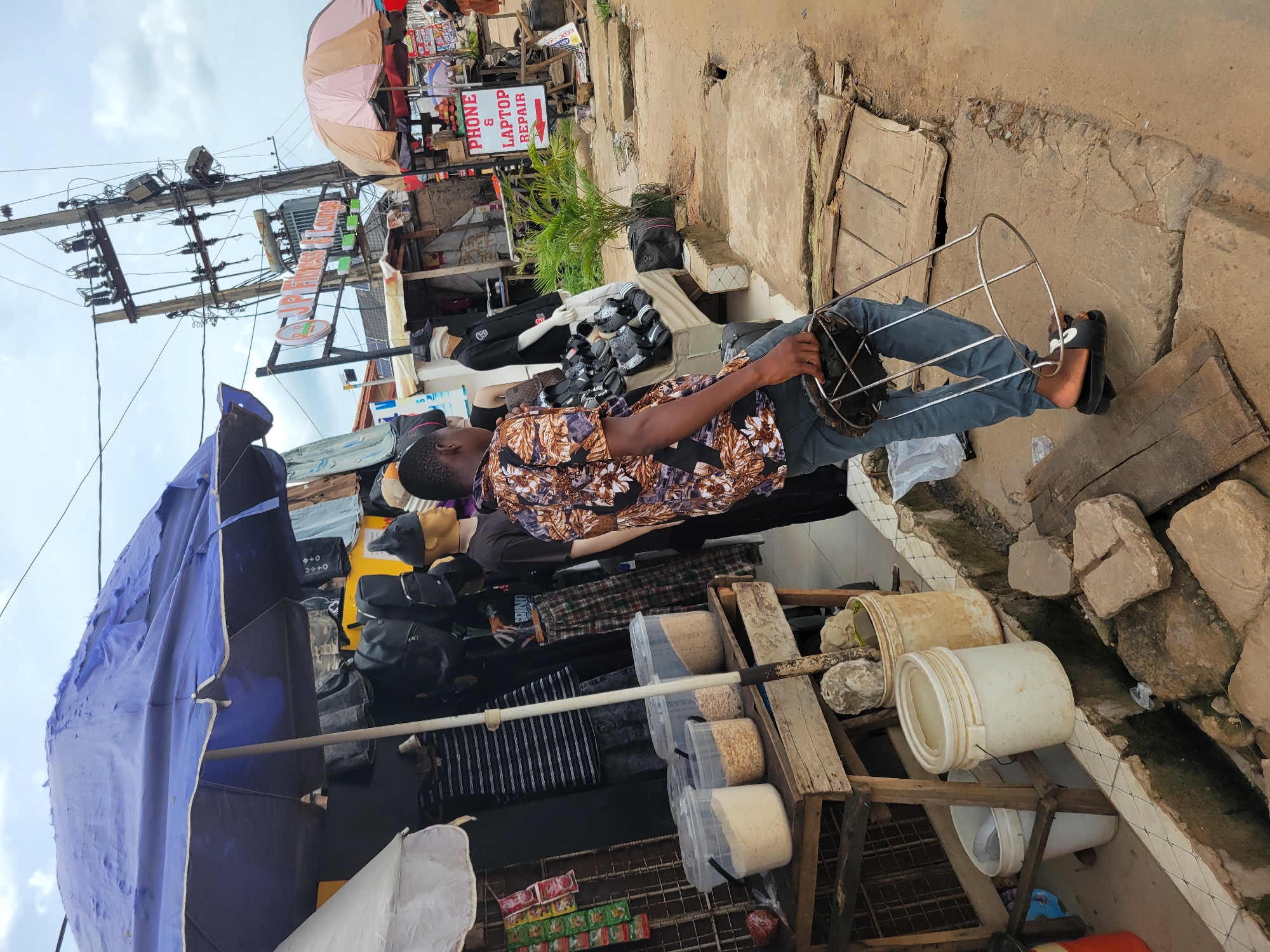 The image depicts a street scene in Egbagu, Nigeria. A man in a patterned shirt and jeans stands on a sidewalk, holding a metal frame. To his left is a market stall with a blue umbrella, displaying items such as bags, clothing, and containers of grains. Behind the stall are mannequins and advertisements. The backdrop includes buildings, utility poles, and a cloudy sky. The ground consists of concrete and assorted debris.