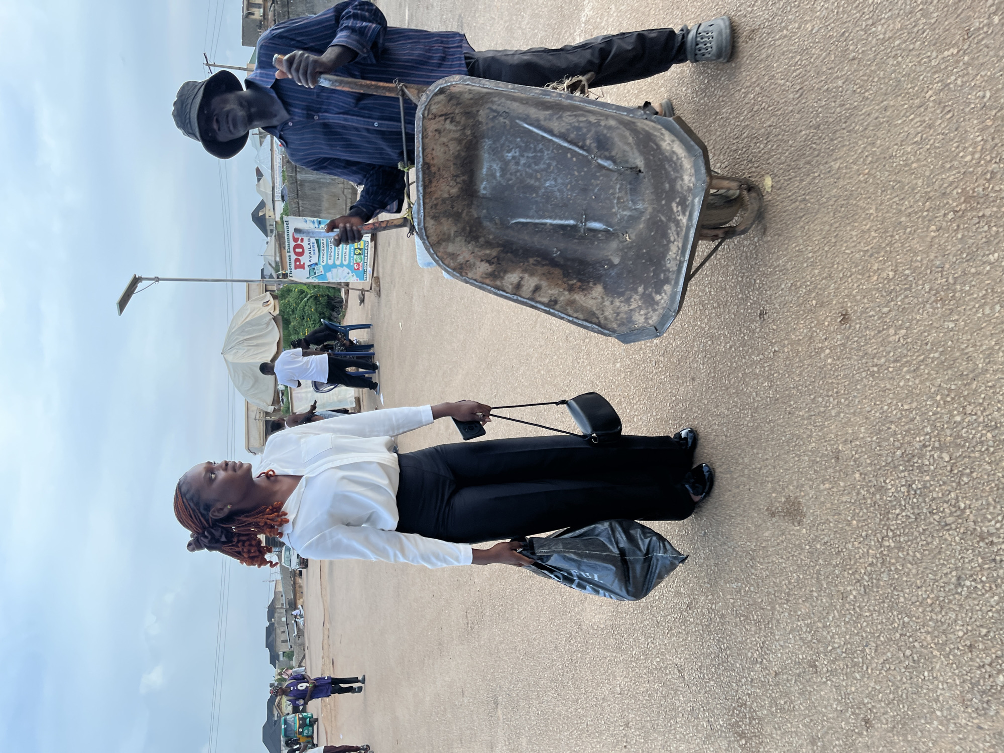 This outdoor street scene in Rafin Par, Nigeria, features two individuals against a backdrop of buildings and an overcast sky. The ground is a light-colored, textured pavement.
In the foreground, a dark-skinned man, wearing a dark blue striped shirt, dark trousers, and a dark bucket hat, is holding a weathered, dark grey metal wheelbarrow. His left foot, clad in a grey Croc-style shoe, is visible near the wheelbarrow. Adjacent to him, a dark-skinned woman with reddish-brown braided hair, styled upwards, stands in a white long-sleeved shirt and black trousers. She is carrying a small black handbag over her shoulder, holding a black plastic bag, and a black smartphone in her right hand, and is looking upwards and slightly to her left.
The background shows various low-rise buildings and scattered individuals, some seated under a white umbrella. A street light with a solar panel is prominent on the left side of the frame. A significant detail is a light blue and white signboard, displaying the text "Thomas Emmanuel," "POS," "AVAILABLE HERE," "WITHDRAWAL," "DEPOSIT YCM," and "OPEN ACC," indicating a point-of-sale service. The scene depicts everyday activity on a cloudy day.