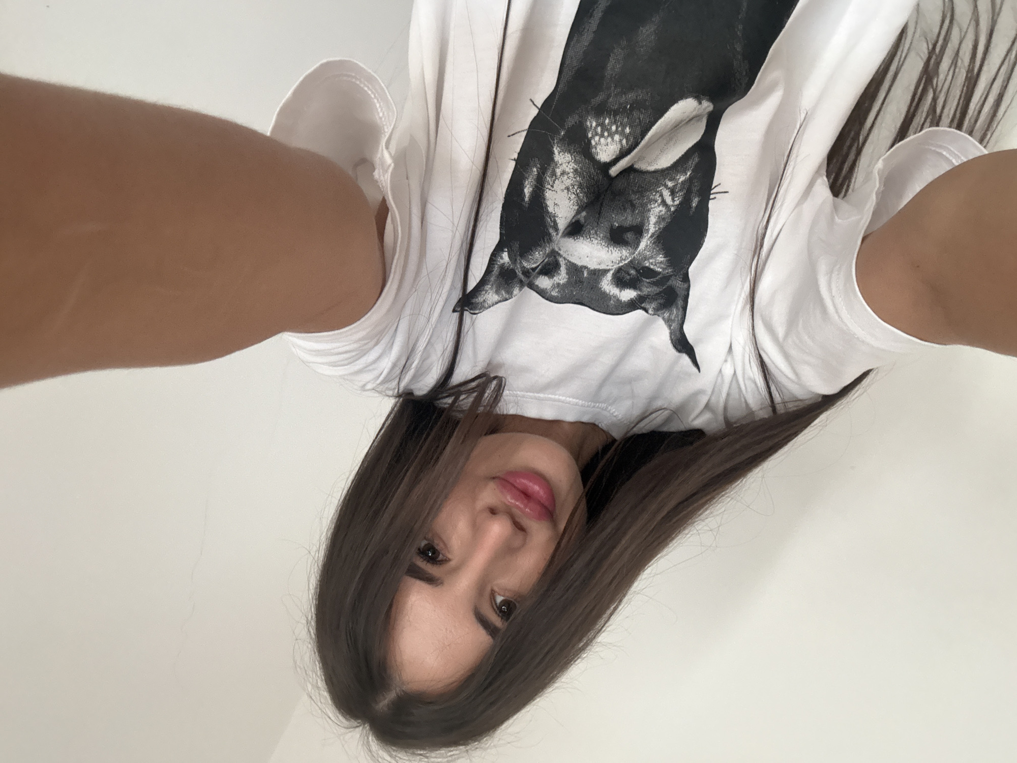 A young woman with long, dark hair and plump lips is taking a selfie. She is wearing a white t-shirt with a black and white graphic of a dog's face printed on it. The background is a plain, white wall. The image is taken from a low angle, with the woman's arm extended towards the camera. There is no visible indication of the location being Chișinău, Moldova.