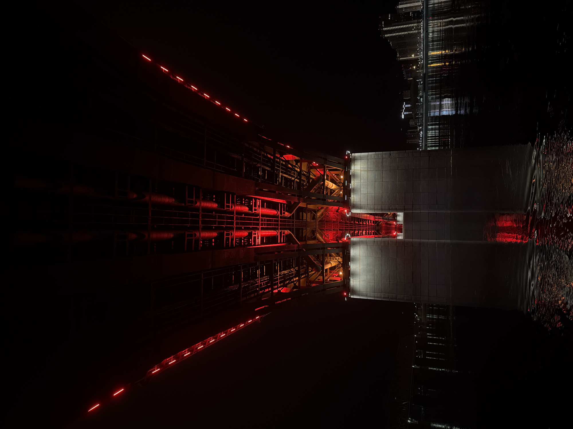 The image shows a bridge, likely in Krasnoyarsk, Russia, at night, with a reflection in the water below. The bridge is illuminated with red lights. Some buildings on the other side of the river are also reflected in the water. The overall scene is dark.