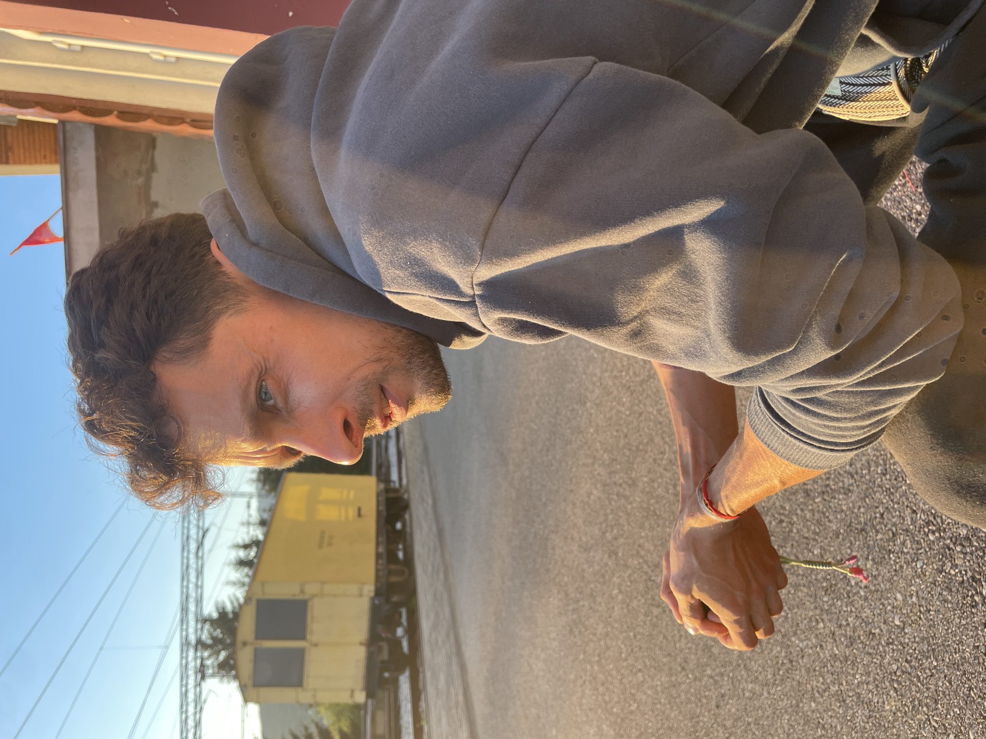 A medium shot photograph captures a man wearing a grey hooded sweatshirt from the shoulders up. He is angled downward, looking left with his gaze directed toward the ground. His hair is dark and curly, and he has a light beard. His right arm is visible, showing the veins, and he wears a red bracelet. In his hand, he holds a small flower. Behind him is a railway track with a yellow cargo train, under a bright sky. In the background, on the left, is a building with a red flag on its roof. The image appears to be taken on a street in Mojkovac, Montenegro.