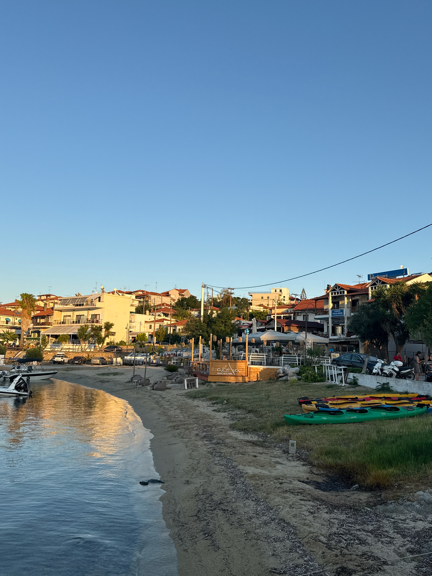 The image depicts a sunlit scene along the waterfront of Ammouliani, Greece. In the foreground, calm, blue water reflects the golden light of the setting sun. The shore is a sandy beach scattered with dried seaweed. To the right of the beach, several brightly colored kayaks are neatly stacked on the grassy embankment. Behind the beach, a row of buildings line the waterfront, featuring white and beige facades with terracotta roofs, characteristic of Mediterranean architecture. Some buildings house cafes and restaurants with outdoor seating areas and umbrellas. Several parked cars and motorcycles are visible along the street behind the cafes. A few people are seen walking and standing near the establishments. In the background, the town extends up a gentle slope, with more buildings and greenery. The sky is a clear, pale blue.