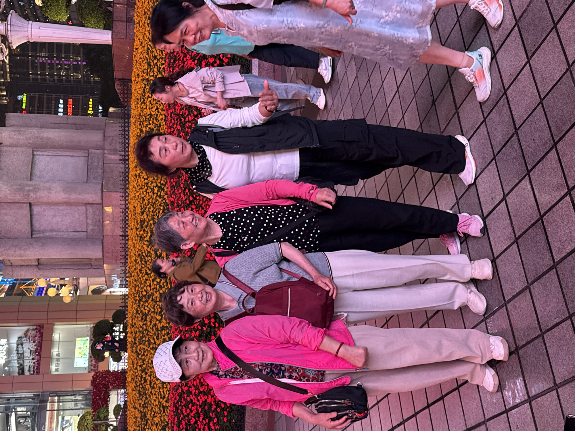 The image shows a group of approximately six elderly women posing for a photo in what appears to be a public square in Yuzhong District, China. They are positioned in a line, with each woman dressed in a distinct outfit. The background includes a flower bed with red and orange blossoms. Buildings are visible in the background, suggesting an urban setting. The women are standing on a brick walkway. The image appears to have been taken in the evening or at night, based on the lighting conditions.