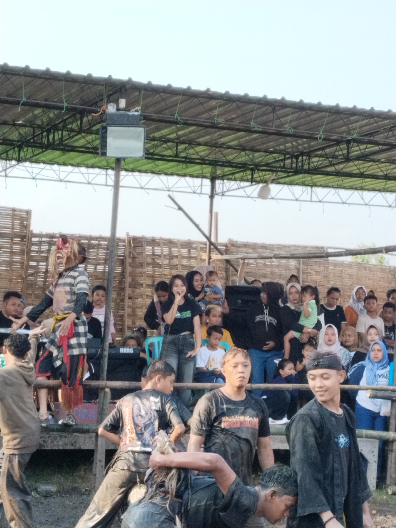 This image captures a vibrant outdoor cultural event in Kedawung, Indonesia, featuring a traditional performance for a gathered community.
In the foreground, several young men are visible, some covered in mud or dirt, suggesting active participation in a physical event. One man, seen from the back, wears a dark t-shirt with a graphic design. Another man, facing the viewer, has a concentrated expression.
The midground features the central performance area. A key performer, possibly a dancer or character, stands on a raised wooden platform. This individual wears a striking, elaborate mask with red and white features, sharp fangs, and a flowing wig, accompanied by a striped shirt, black pants, and a red and black checkered sarong-like garment. To the performer's right, a woman sings passionately into a microphone, positioned near a large black speaker. Behind her, a "KORG" brand keyboard is visible, suggesting live musical accompaniment.
An elevated, tiered seating area constructed from bamboo and wood holds a diverse audience, comprising adults and children. Many spectators are intently watching the performance, with some smiling and holding infants or young children. The audience appears engaged and entertained.
The setting is outdoors, beneath a corrugated metal roof supported by a metal truss structure, providing shade. A large floodlight is mounted to this overhead structure. A rustic bamboo lattice wall serves as a backdrop to the performance area. The ground in the immediate foreground appears muddy or wet, indicating the nature of the activity.
The time of day appears to be daytime, likely late afternoon, under a clear sky. The overall atmosphere is lively and communal, indicative of a local celebration or cultural display.