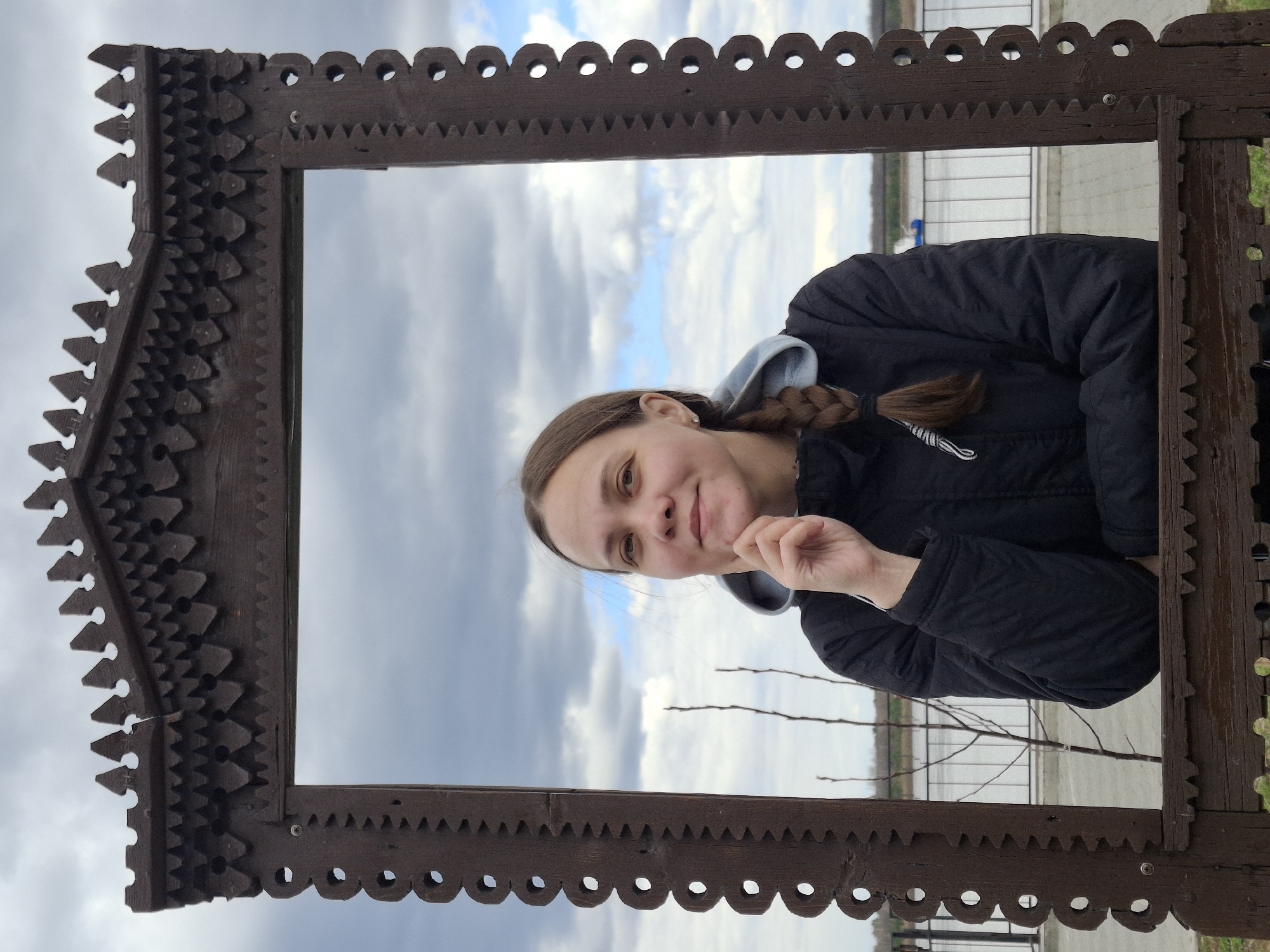 A young woman is depicted in a vertical portrait, framed by an elaborate, dark brown wooden structure. The woman stands centrally within the frame, looking directly towards the viewer with a gentle, pleasant smile. Her light brown hair is styled in a braid that rests over her left shoulder. She is dressed in a dark, possibly puffer, jacket worn over a light-colored hooded sweatshirt, the hood of which is visible around her neck. Her right hand is raised to her chin, suggesting a thoughtful or relaxed pose.
The wooden frame itself is highly ornate, featuring intricate carvings. The inner edges have a scalloped design with small circular cutouts, while the outer perimeter, particularly the corners, displays sharp, jagged, and layered patterns, characteristic of traditional folk art or architectural detailing.
The background reveals an outdoor setting under a partly cloudy sky, with hints of blue visible between the grey clouds. Behind the woman, a calm body of water, likely a river, extends horizontally, with indistinct land or structures visible on the distant bank. A few bare tree branches are visible on the right side, just within the frame's opening. The diffused lighting suggests an overcast day. This scene is set in Sarapul, Russia. No text is visible in the image.