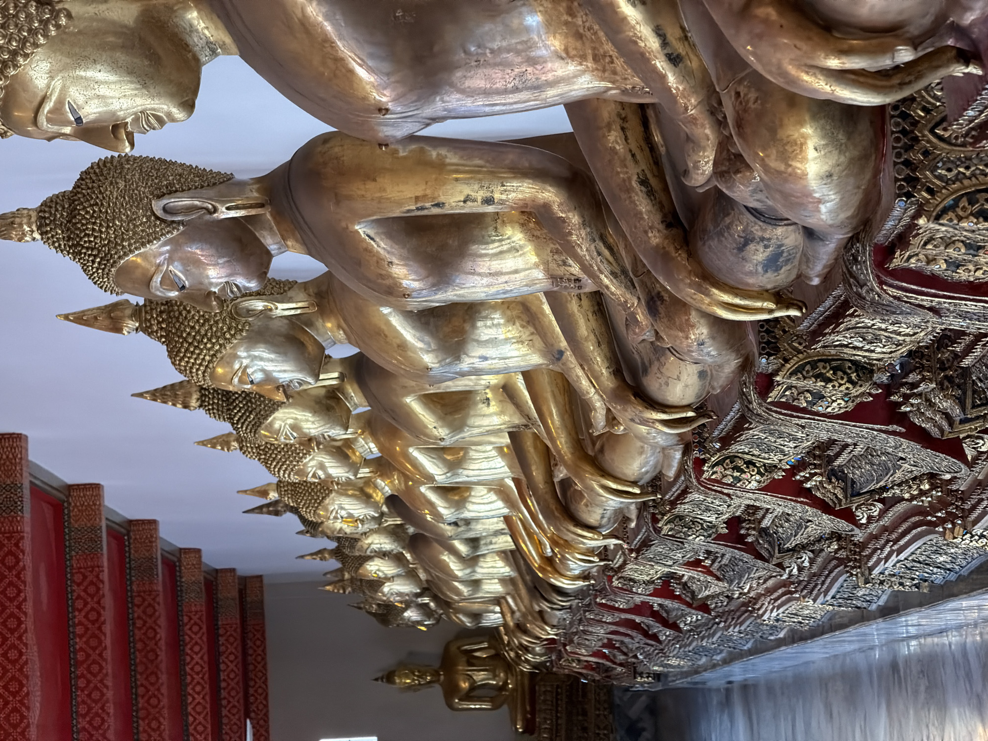 The image depicts a row of golden Buddha statues lined up in a temple in Bangkok, Thailand. The statues are seated in a meditative pose, with their hands resting on their laps. The foreground of the image is dominated by the statues, which are highly detailed and appear to be made of polished brass. The statues are adorned with elaborate decorative elements, including intricate carvings and patterns. In the background, a red ceiling with intricate golden patterns can be seen, suggesting the grandeur of the temple. The overall impression is one of serenity and reverence.
