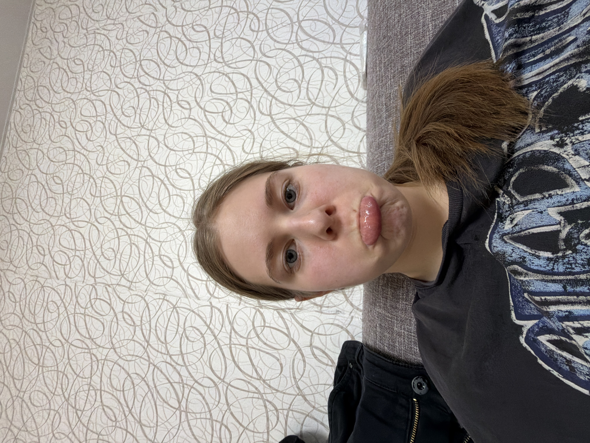 An indoors portrait captures a young woman posing playfully for the camera. The image is oriented sideways, with her head to the left and her body extending to the right, suggesting she is lying on a sofa or chair. She has fair skin, light blue eyes, and light brown hair tied back in a ponytail, which rests on her right shoulder. Her facial expression is a distinctive pout, with her lips pursed forward and a glimpse of her tongue visible, conveying a silly or humorous mood.
She is wearing a dark, possibly black, t-shirt adorned with a large, stylized graphic design on the right side of her chest and shoulder. The design features intricate, distressed patterns in shades of light blue and white, resembling the head of an animal or mythical creature. Below her t-shirt, the top of dark pants, possibly jeans, with a visible button and zipper, can be seen.
The background consists of a light-colored wall (off-white or light beige) covered with a repeating pattern of swirling, interconnected lines or loops in a slightly darker, muted tone. To her right, a textured fabric surface, likely part of a sofa or armchair, is visible in a dark grey or brown color. No specific time of day or outdoor weather conditions can be inferred, and there are no discernible location cues within the image itself to confirm the provided context of Perm, Russia. No visible text is present.