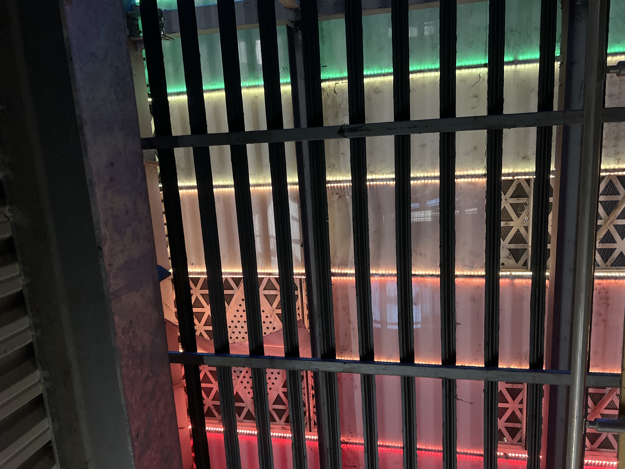 The image depicts a view looking up through a metal grid or bars, possibly a ceiling or a security structure. Behind the grid are vertically oriented panels or segments illuminated with colored LED strips - green at the top, fading through yellow/orange to red at the bottom. Some panels have geometric patterns of triangles or cross-bracing behind them. A structural gray support beam is visible on the left side. The location could be related to the architecture and lighting design of Kapana district.