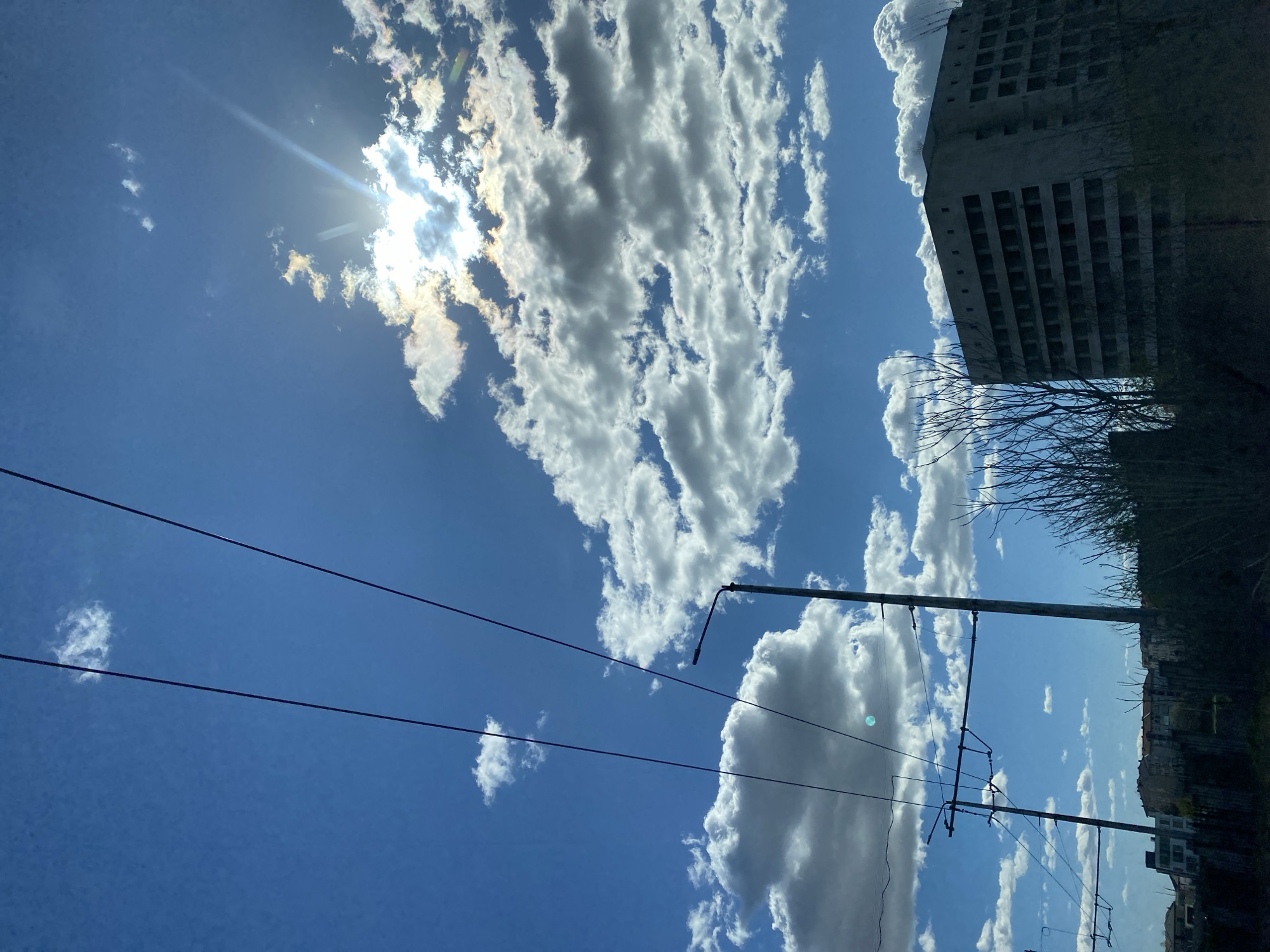 The image shows a bright blue sky with white cumulus clouds, taken in what seems to be the city of Pleven, Bulgaria. A bright sun is visible, partially obscured by clouds, creating sunbeams. Power lines stretch across the sky, connected to utility poles. A large, possibly abandoned, concrete building with multiple window openings is visible on the right side of the image. Some trees and other buildings are also visible.