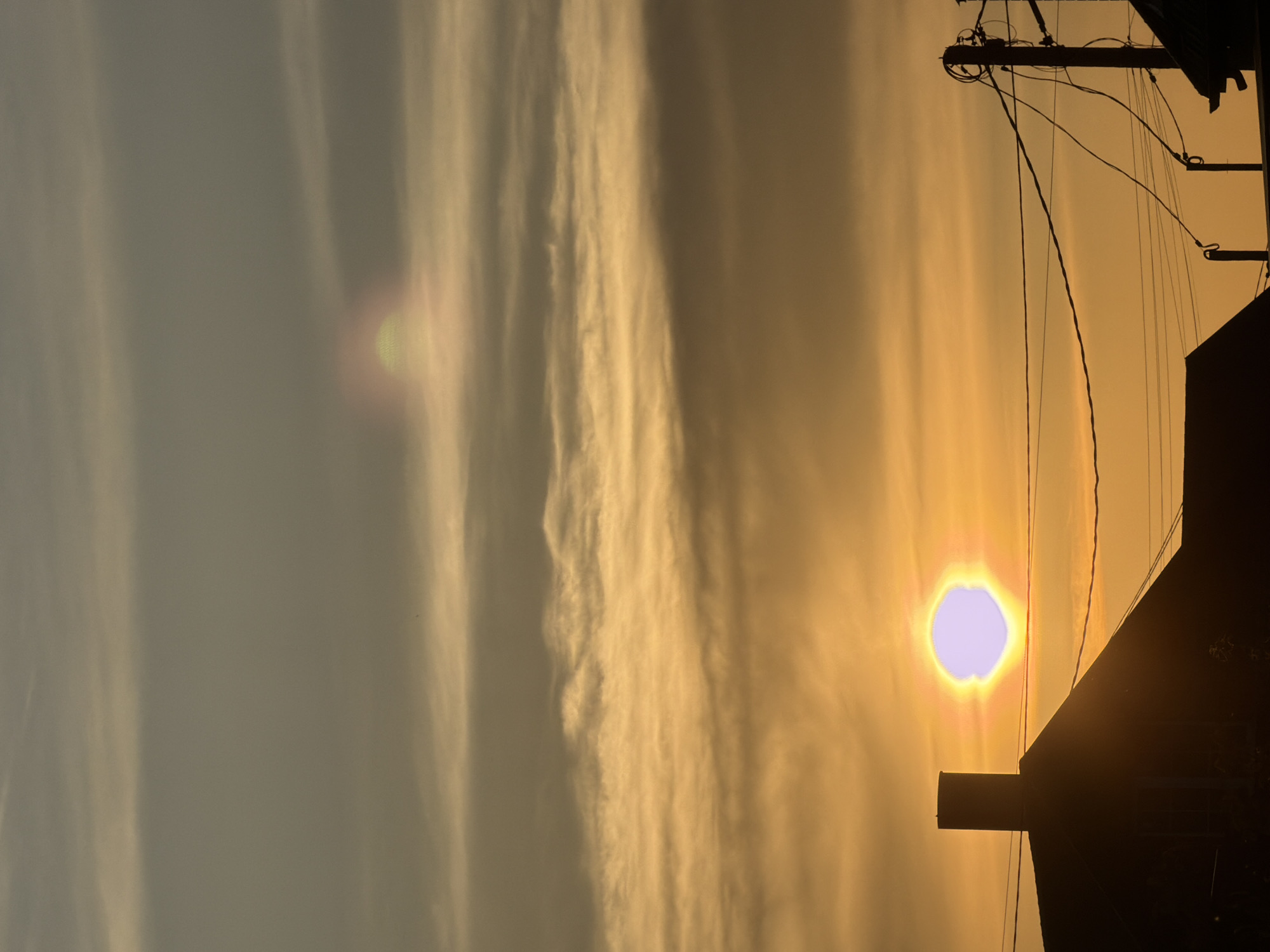 A vertical, outdoor shot taken in Rossiyskiy, Russia. The sky is a gradient of colors, ranging from a pale grey on the left side to a warm golden hue on the right, where the sun is positioned. The sun appears as a bright, circular orb with a faint blue-ish center, radiating yellow and orange light. To the right, the silhouette of a building is visible, with a chimney protruding from the roof. An electric pole is positioned on the right, with wires connecting to the building. Long, streaky clouds stretch across the sky, adding texture to the background.