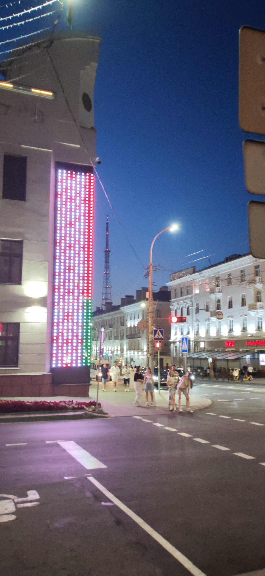 This is a night-time, eye-level shot of a street scene in Homyel, Belarus, likely near a downtown area. On the left, a building facade is illuminated by a tall, vertical LED display showing a pattern of red and white lights, with a vertical stripe of green lights at its right edge. A CCTV camera is visible on the building above the display. In the background, a tall radio tower or transmission tower pierces the deep blue night sky. Across the street, a row of multi-story buildings lines the horizon, many with lit windows and illuminated storefronts, including a prominent KFC sign. A street lamp with a bright light curves over the intersection. Several pedestrians are crossing the street or walking on the sidewalk. They appear to be casually dressed, with some groups talking or looking at their phones. The street itself is paved and marked with white dashed lines indicating a crosswalk. The overall impression is of a lively urban environment at dusk or early evening, with artificial lighting creating a vibrant atmosphere.