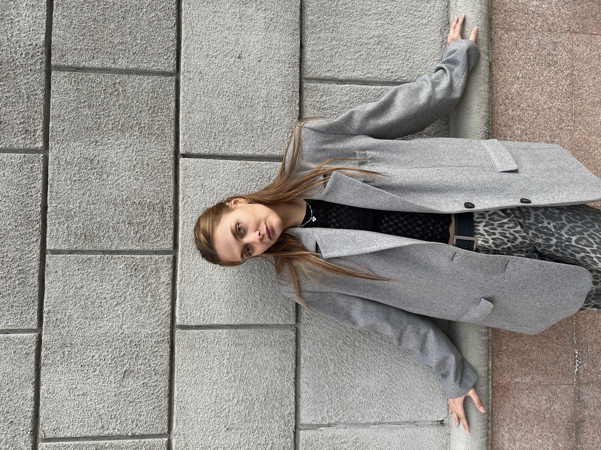 A young woman with long brown hair stands leaning against a stone wall in what appears to be an urban setting. She wears a grey coat over a black top and leopard-print pants, secured with a black belt. Her left arm is extended along the wall, and her right arm is held similarly on another wall to her right. The background features a stone wall made of large rectangular blocks. To the right of the wall is a reddish-brown surface, likely a sidewalk or part of a building.
Without additional information, I am unable to determine the exact location in Krasnoyarsk, Russia.